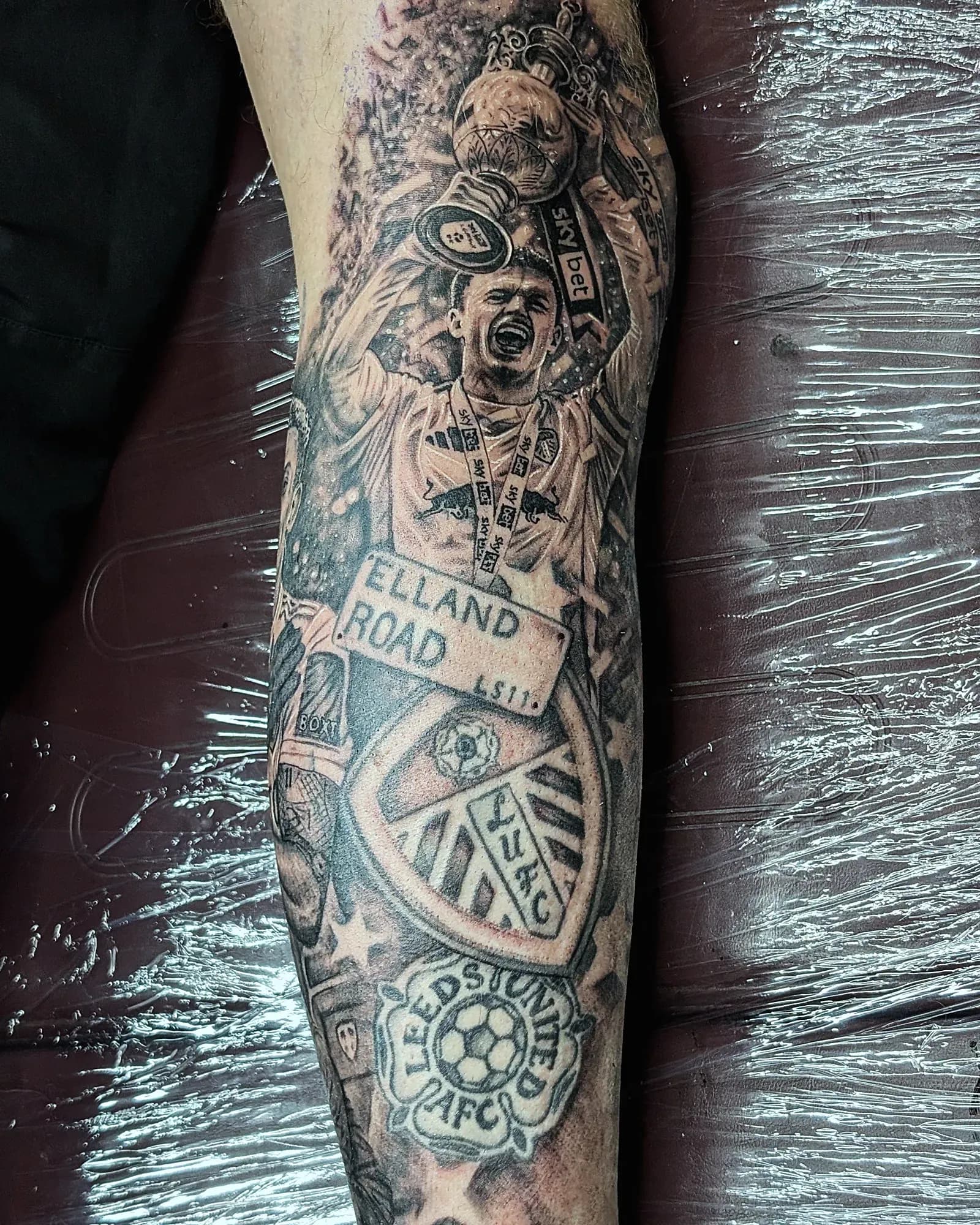 A Leeds United trophy-lift tattoo on the outer calf, fresh from the chair, around an older club badge by another artist.