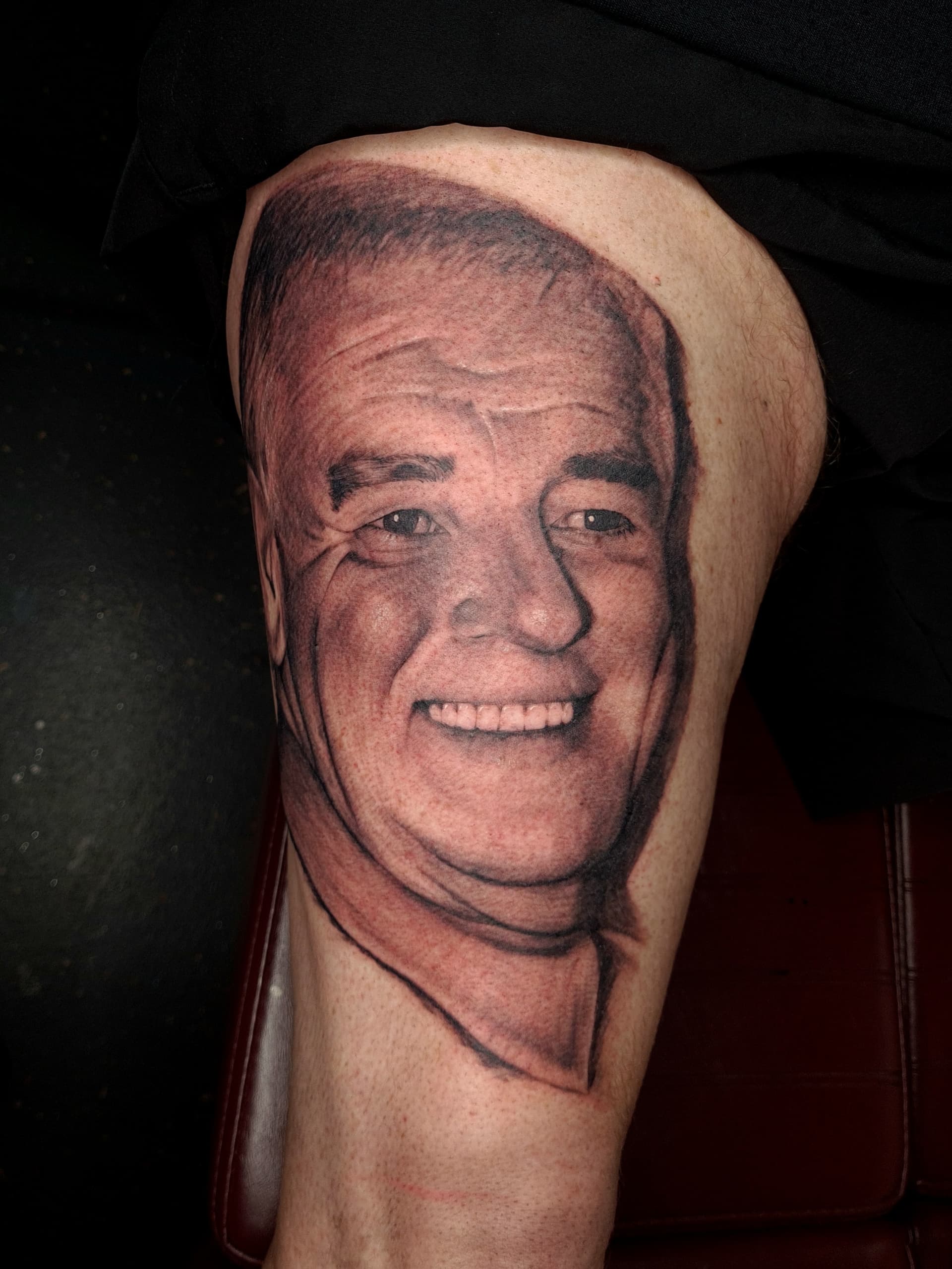 Smiling male portrait tattoo in black and grey realism on leg.