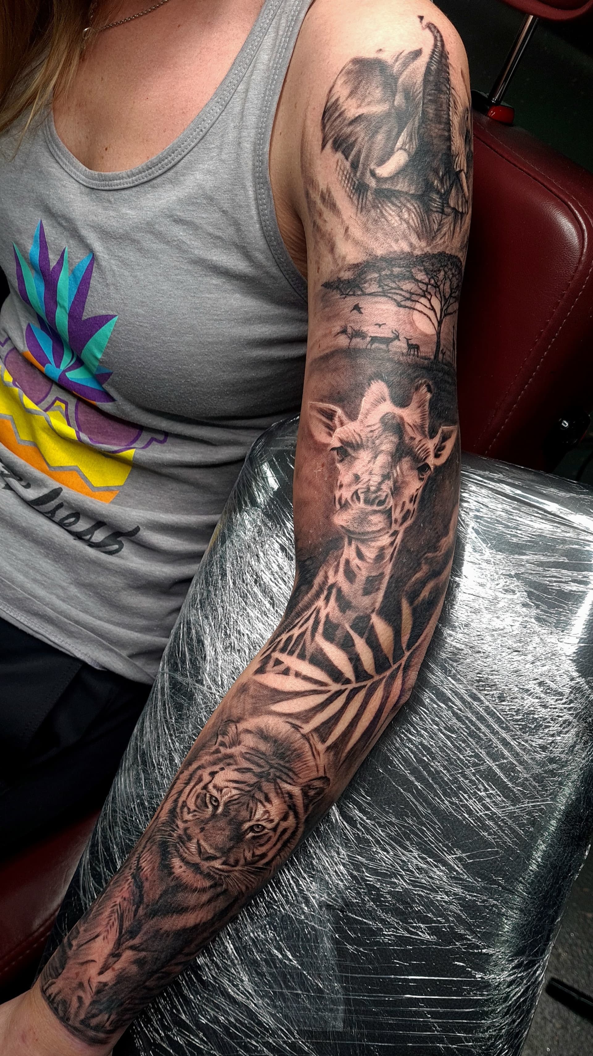 Black and grey realism arm sleeve tattoo featuring an elephant, giraffe, tiger and safari scenery
