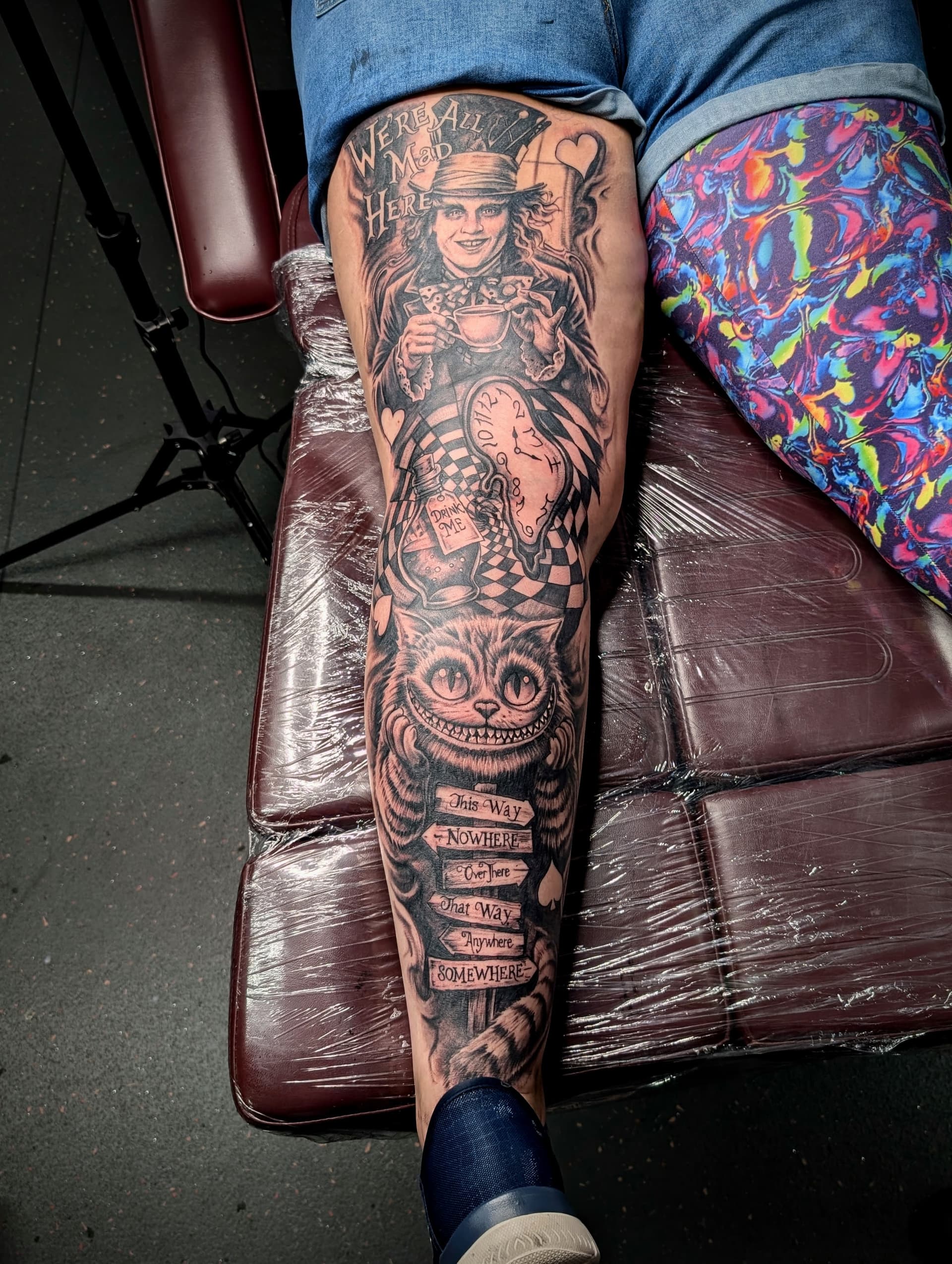 Black and grey realism leg sleeve tattoo of the Mad Hatter, Cheshire Cat and melting clock from Alice in Wonderland