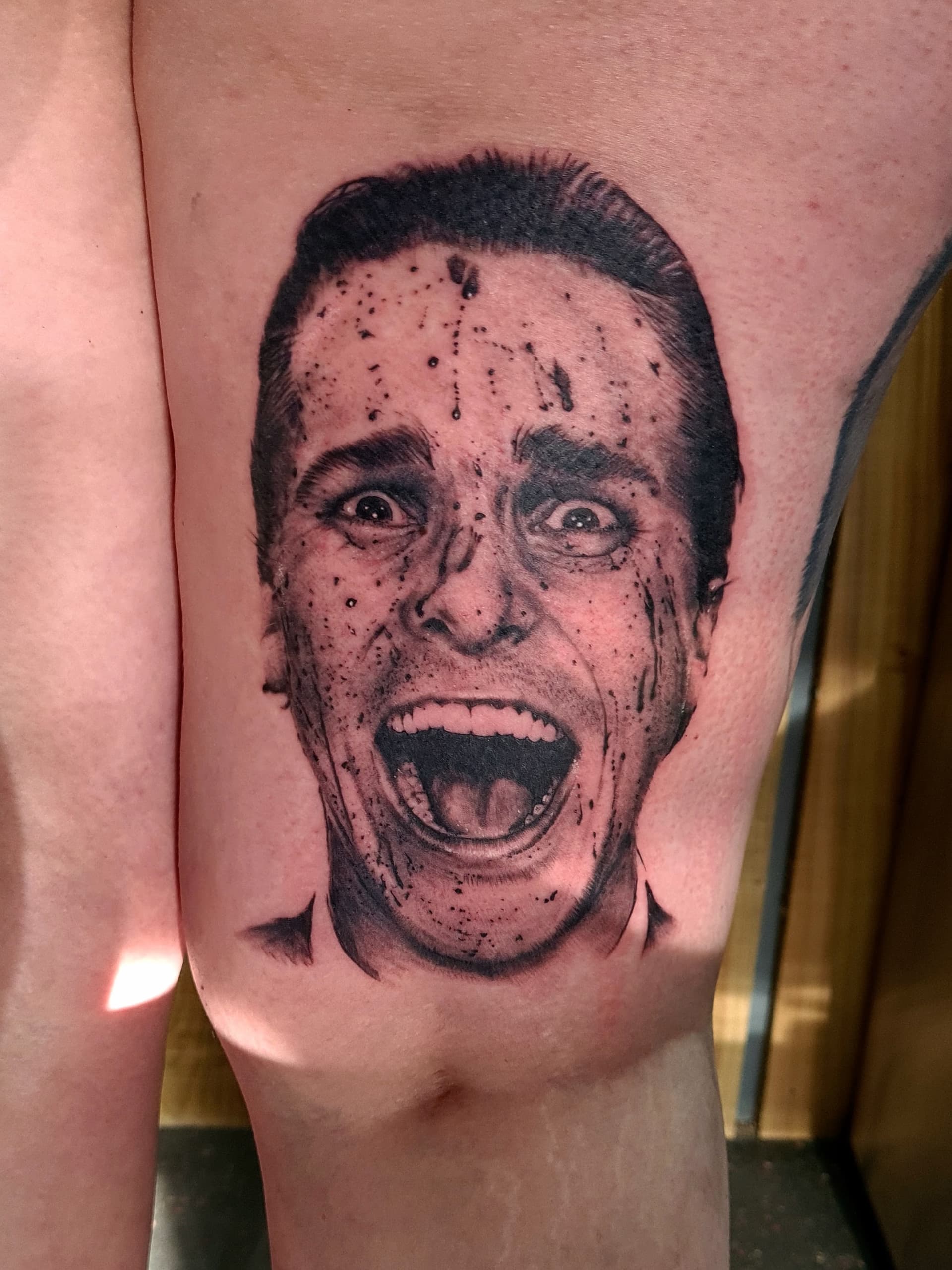 Black and grey realism tattoo of a screaming blood-splattered man's face on the thigh