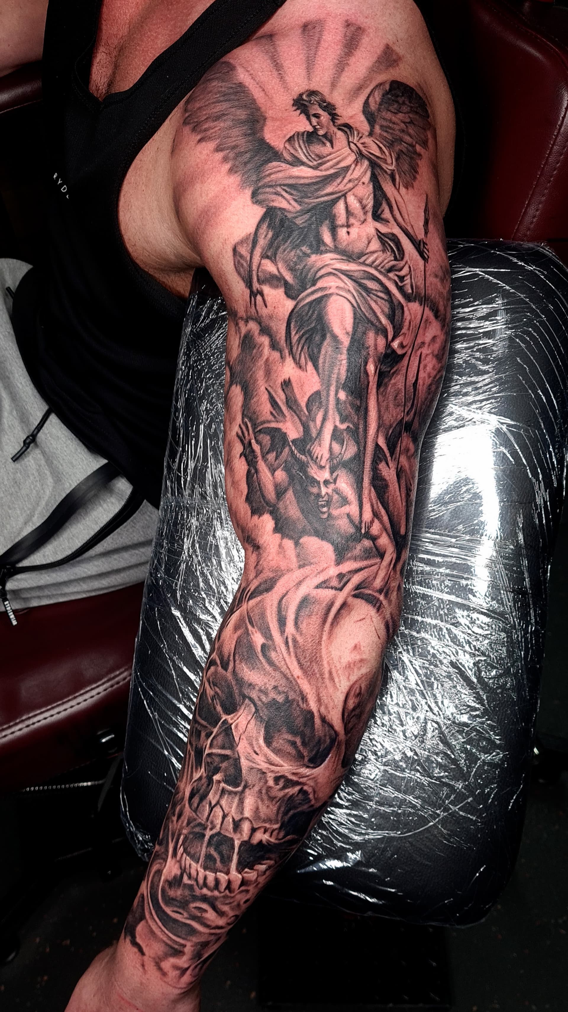 Black and grey realism tattoo of an angel fighting a demon above a skull on the upper arm