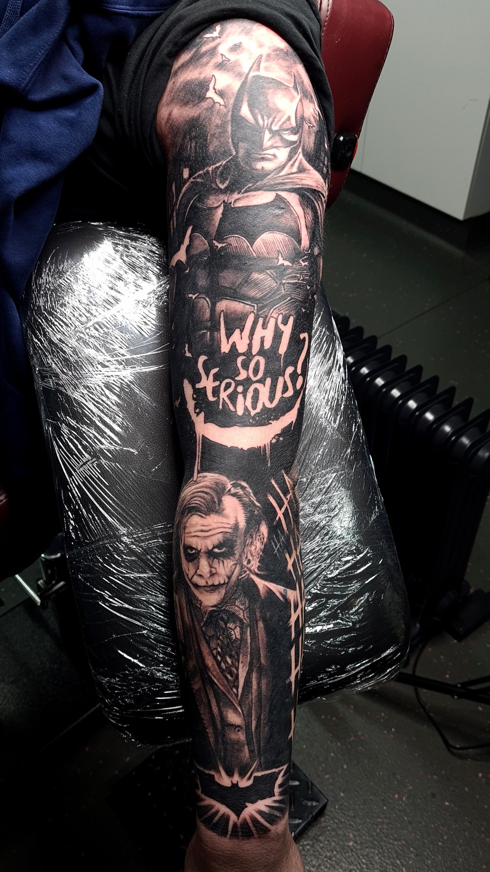 Black and grey realism tattoo of Batman, the Joker and script on the forearm