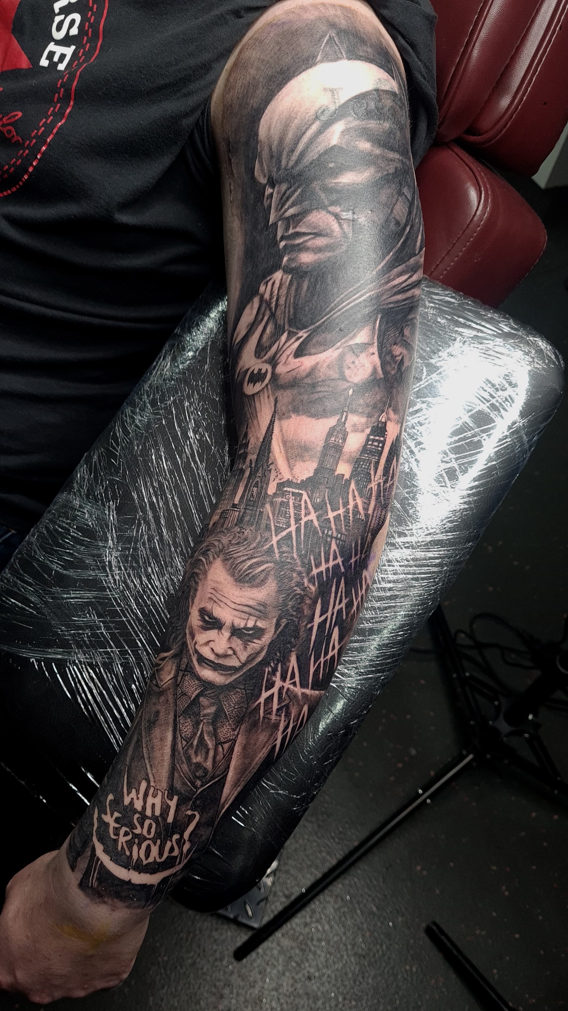 Black and grey realism arm sleeve tattoo featuring Batman, the Joker, and a city skyline