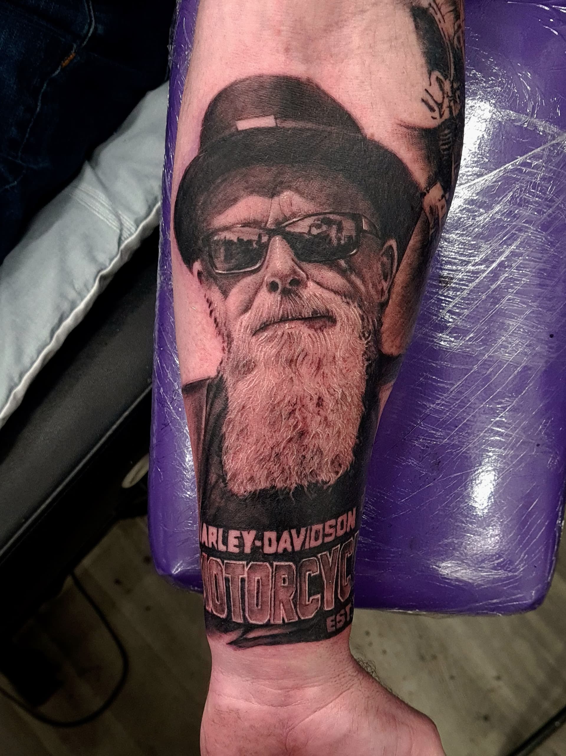 Black and grey realism tattoo of a bearded man in sunglasses and hat with Harley-Davidson script on the forearm