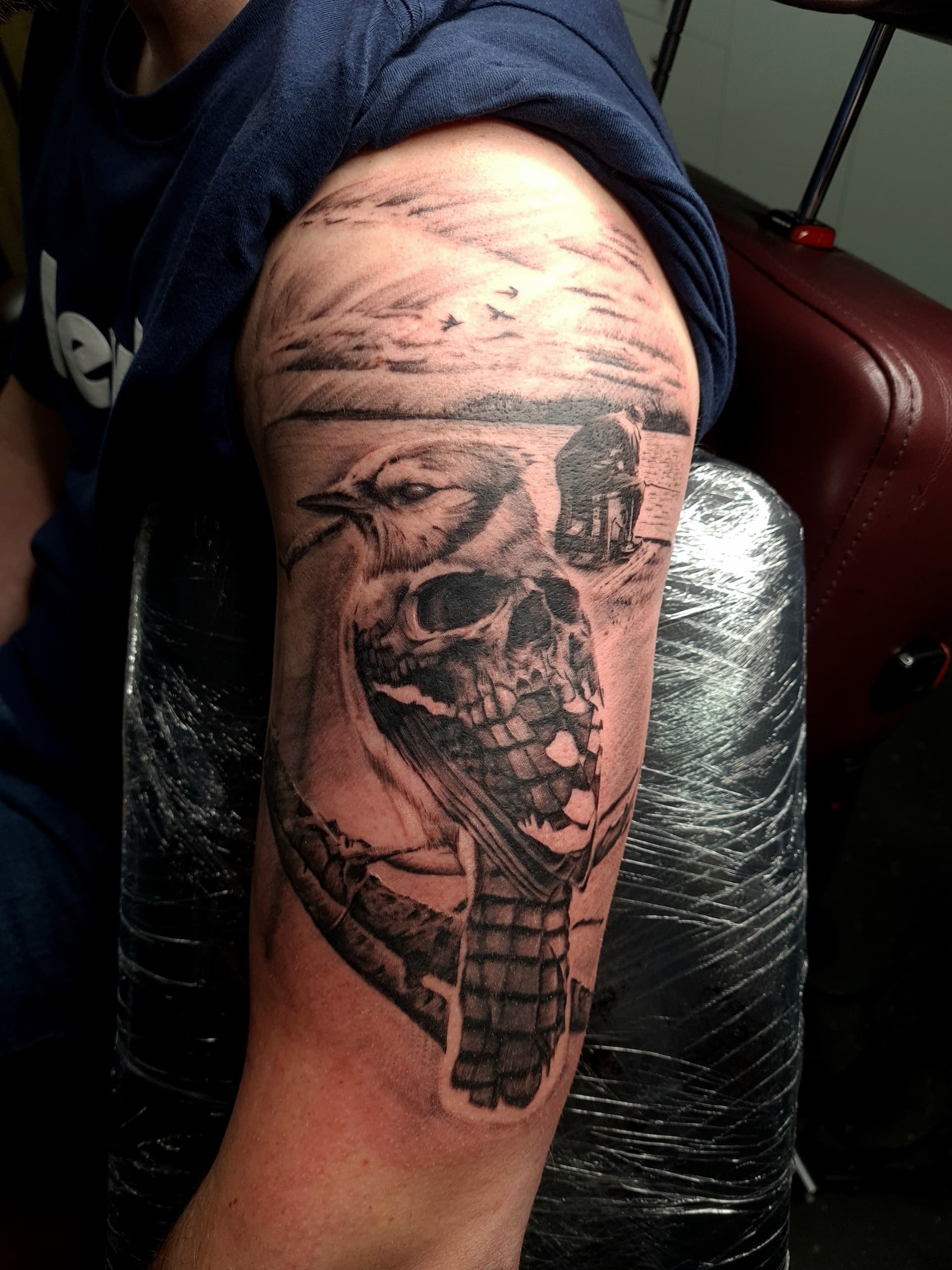 Black and grey realism tattoo of a bird, skull and lakeside scene on the upper arm