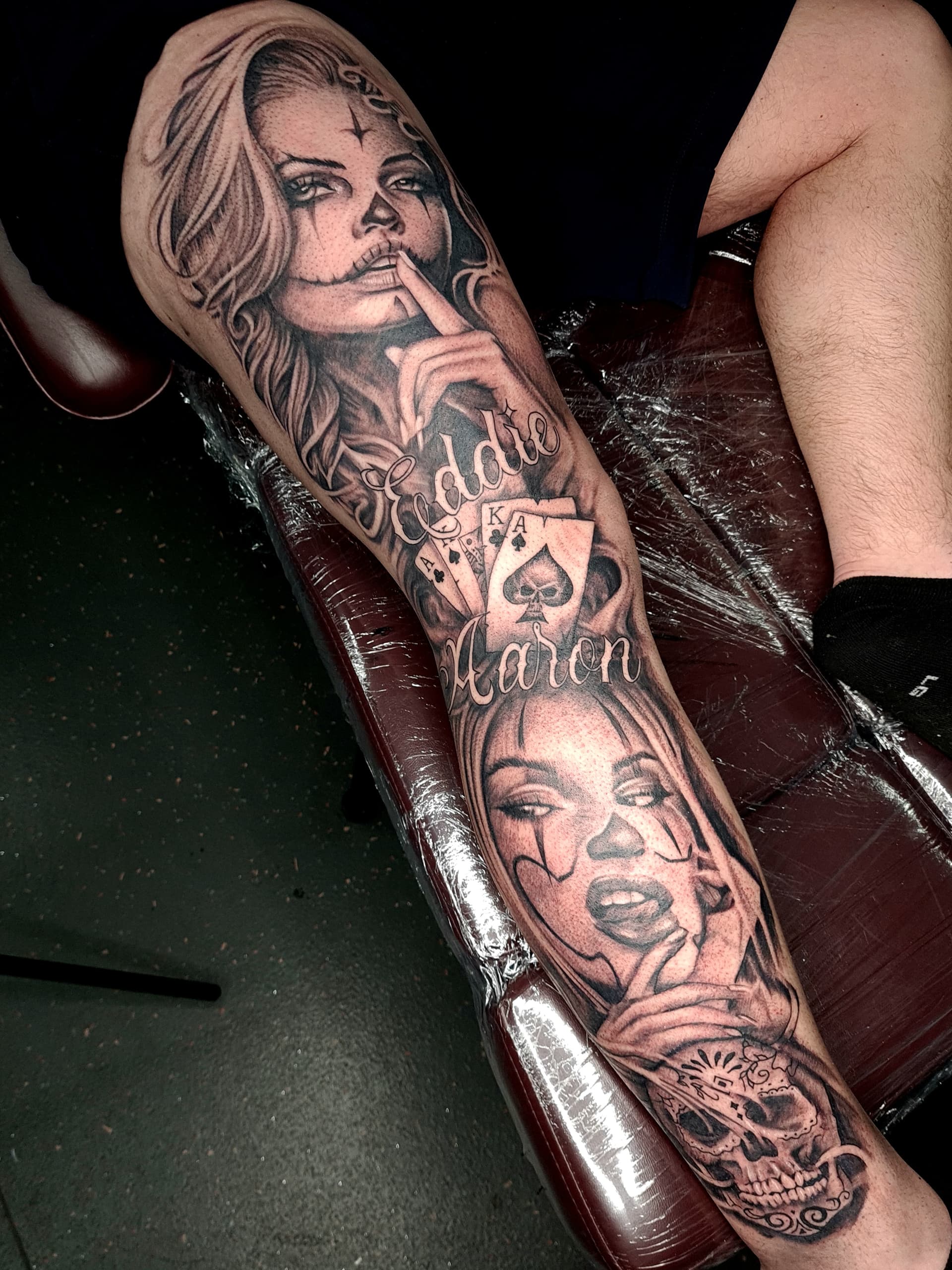 Black and grey realism leg sleeve tattoo featuring two women in clown face paint, playing cards, script and a sugar skull
