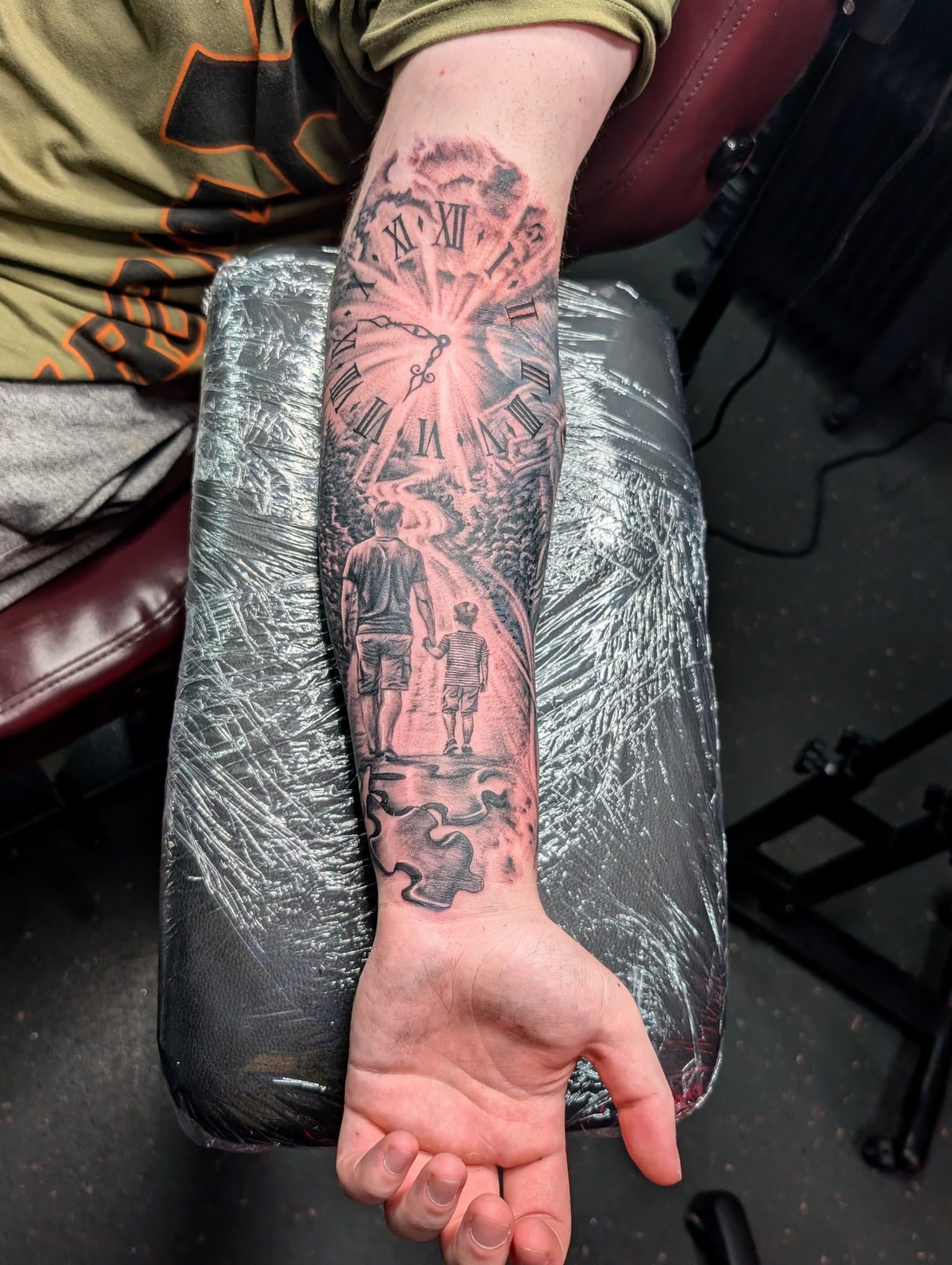 Black and grey realism forearm tattoo of a clock, a path with father and son, and jigsaw pieces