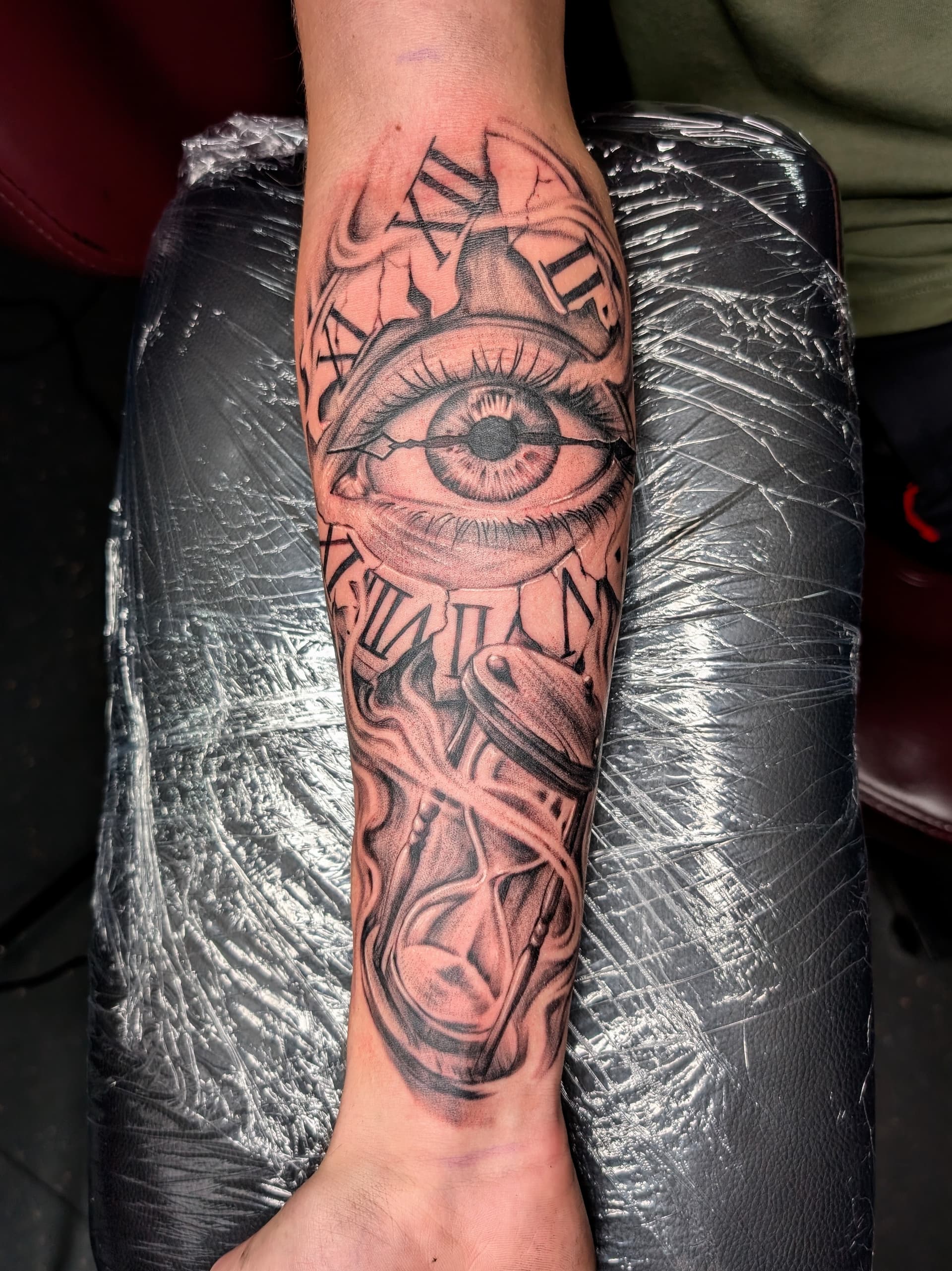 Black and grey realism tattoo of a cracked clock face, eye and hourglass on the forearm