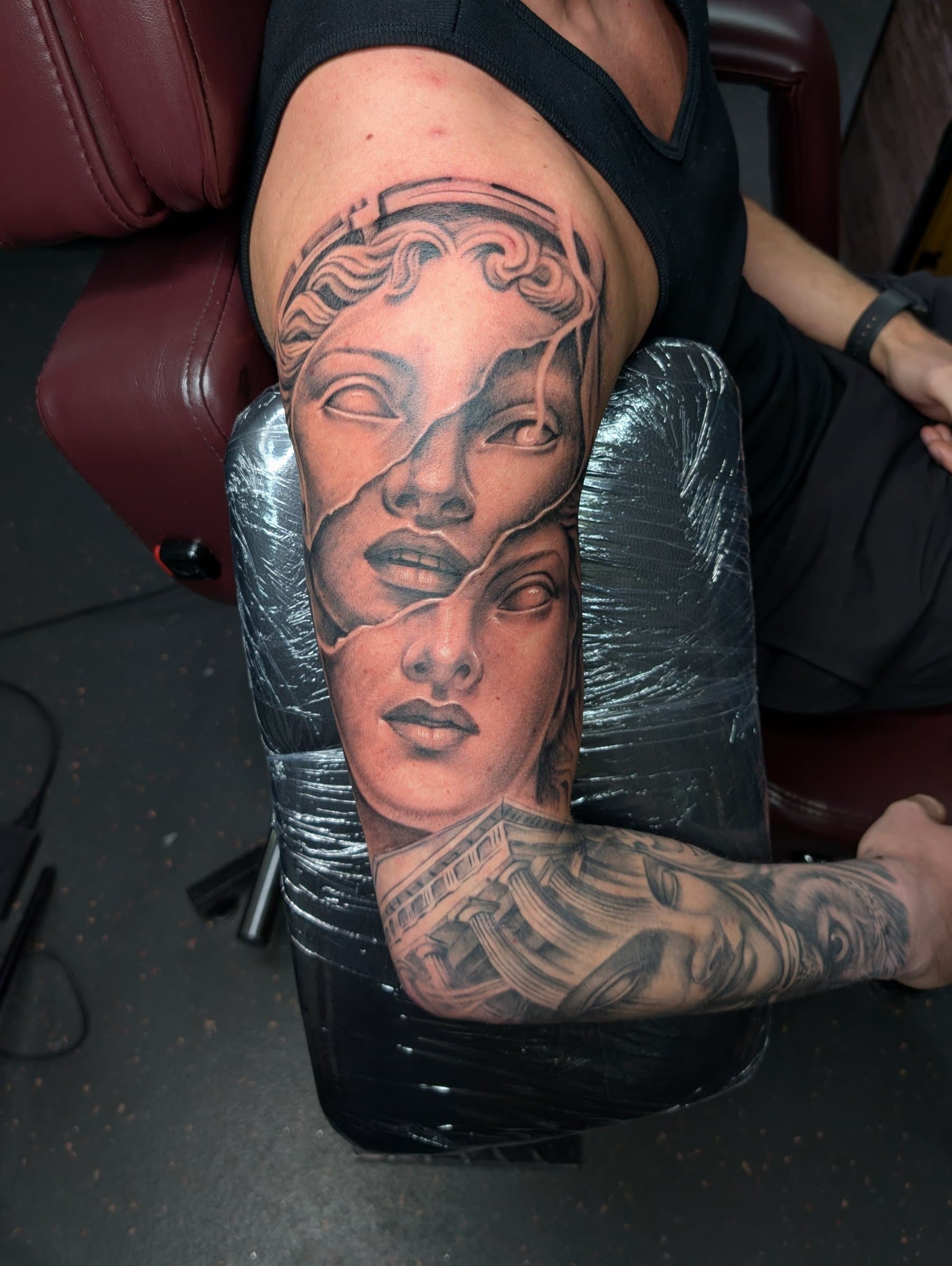Black and grey realism tattoo of layered cracked classical statue faces on the upper arm