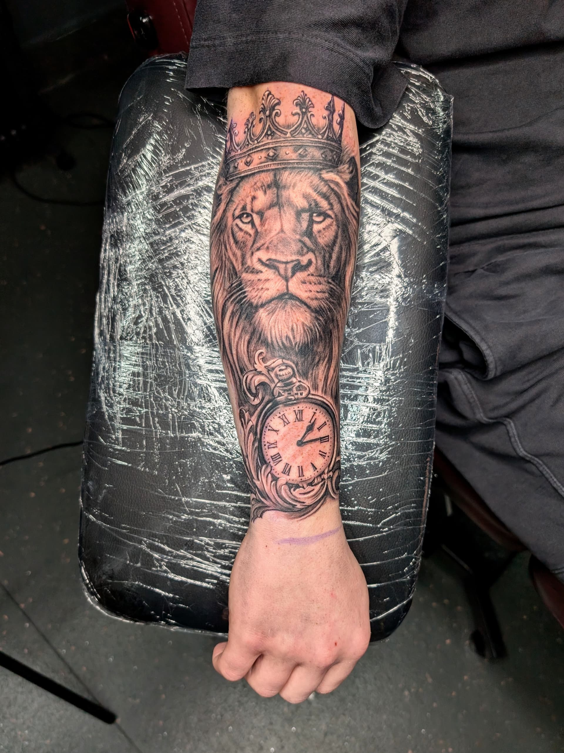 Black and grey realism tattoo of a crowned lion with a pocket watch on the forearm