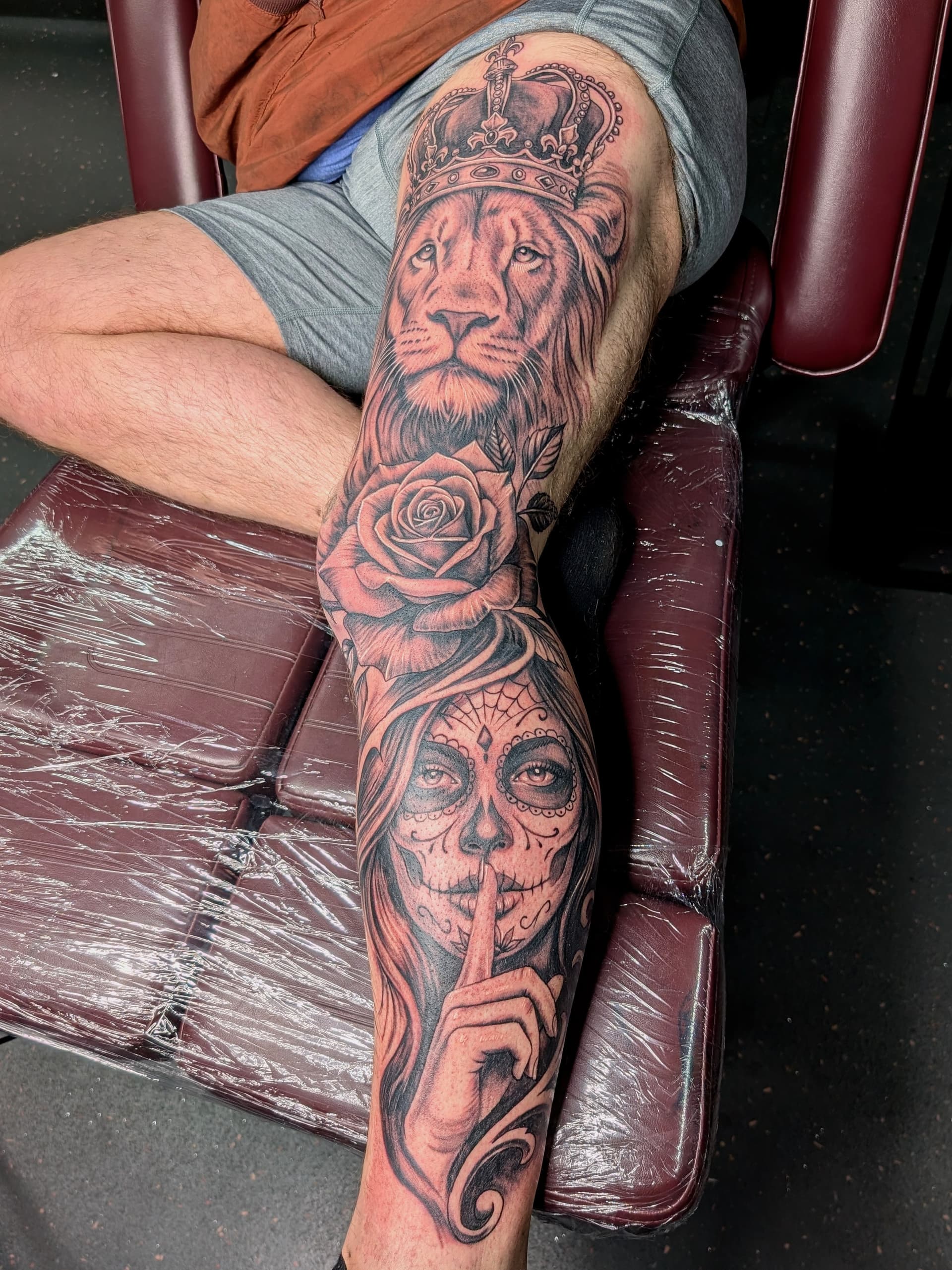 Black and grey realism tattoo of a crowned lion, rose and sugar skull woman covering the leg