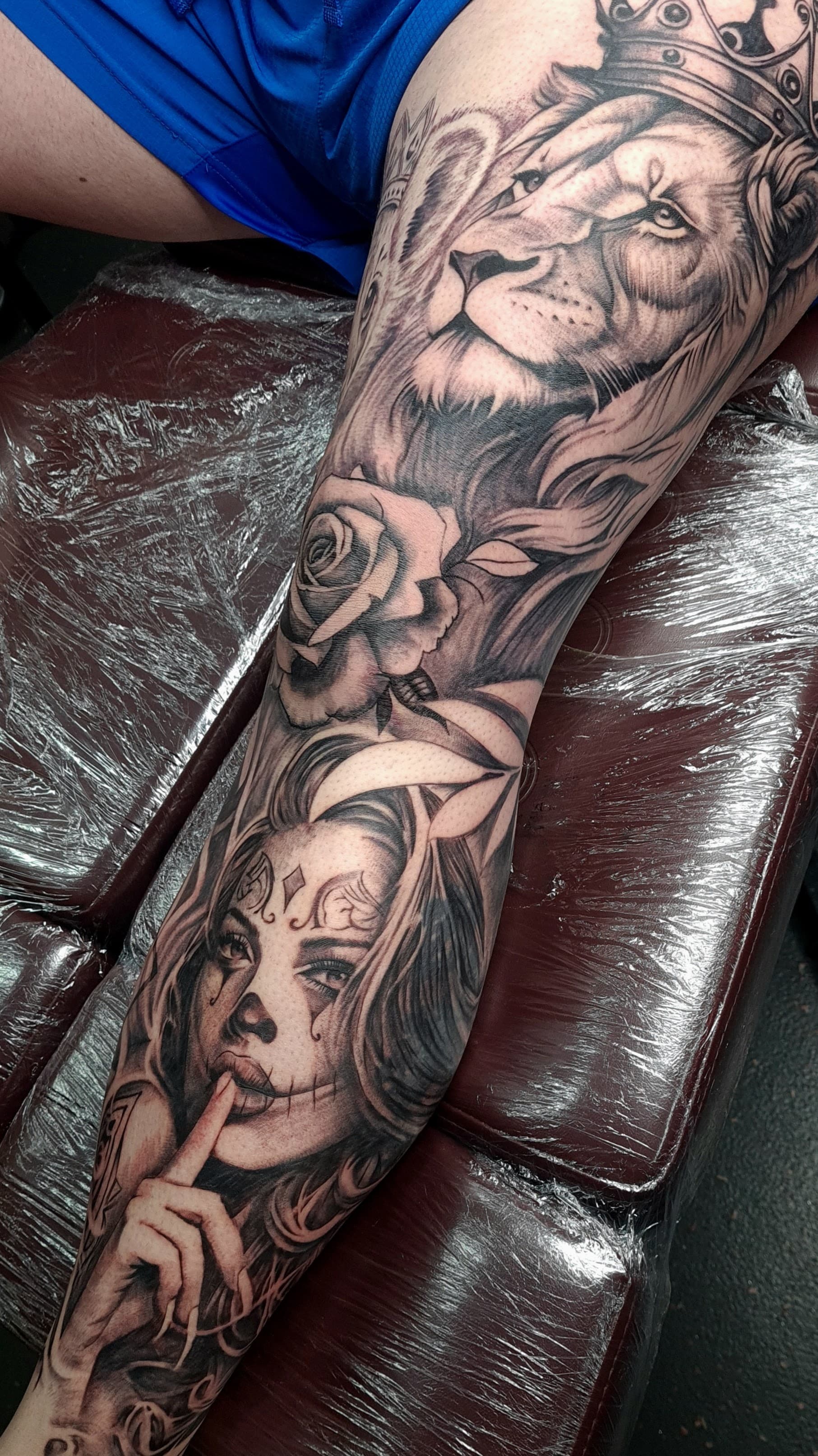 Black and grey realism tattoo of a crowned lion, a rose and a woman with sugar-skull face paint on the thigh