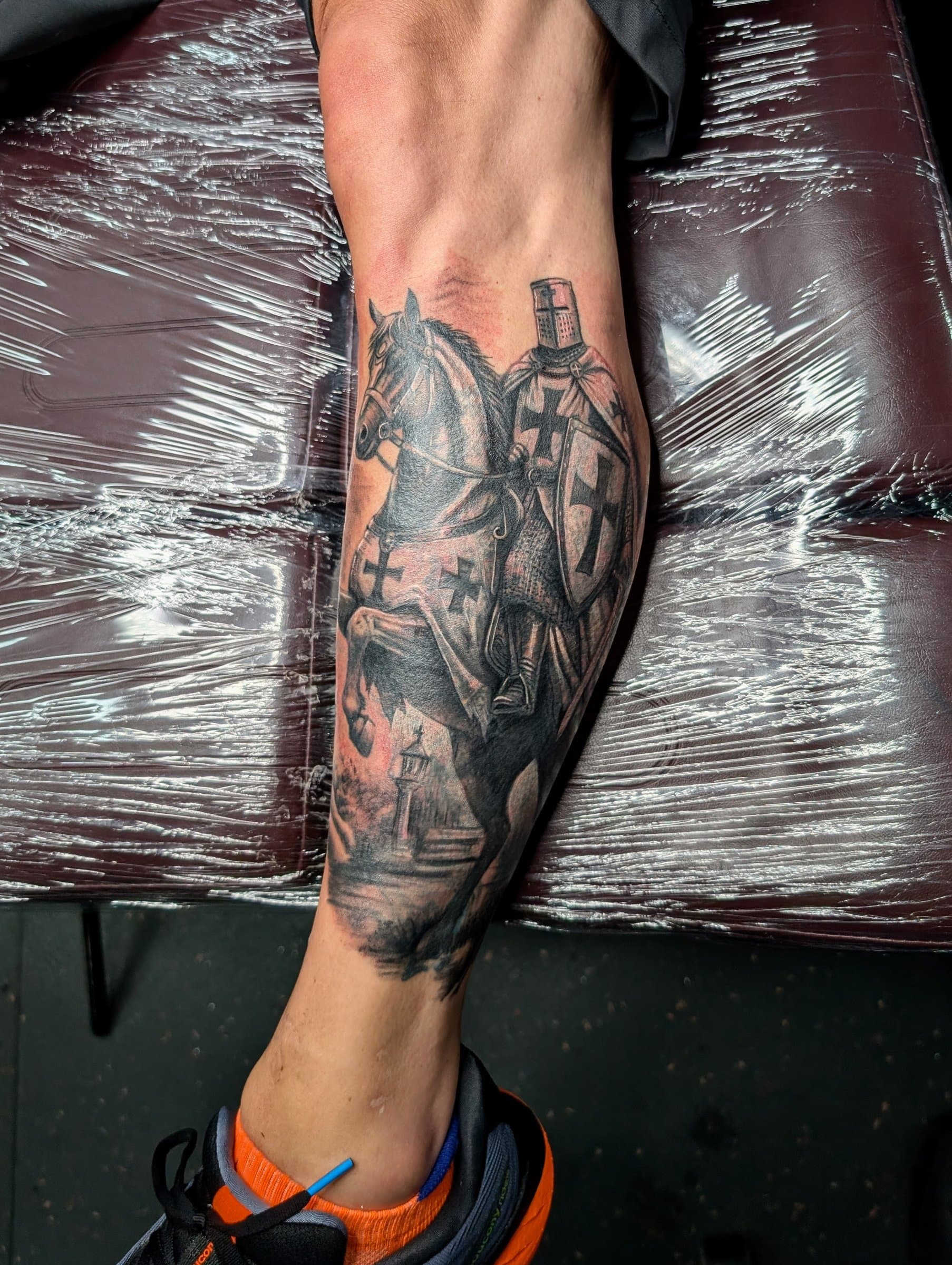 Black and grey realism tattoo of a crusader knight riding a horse on the calf