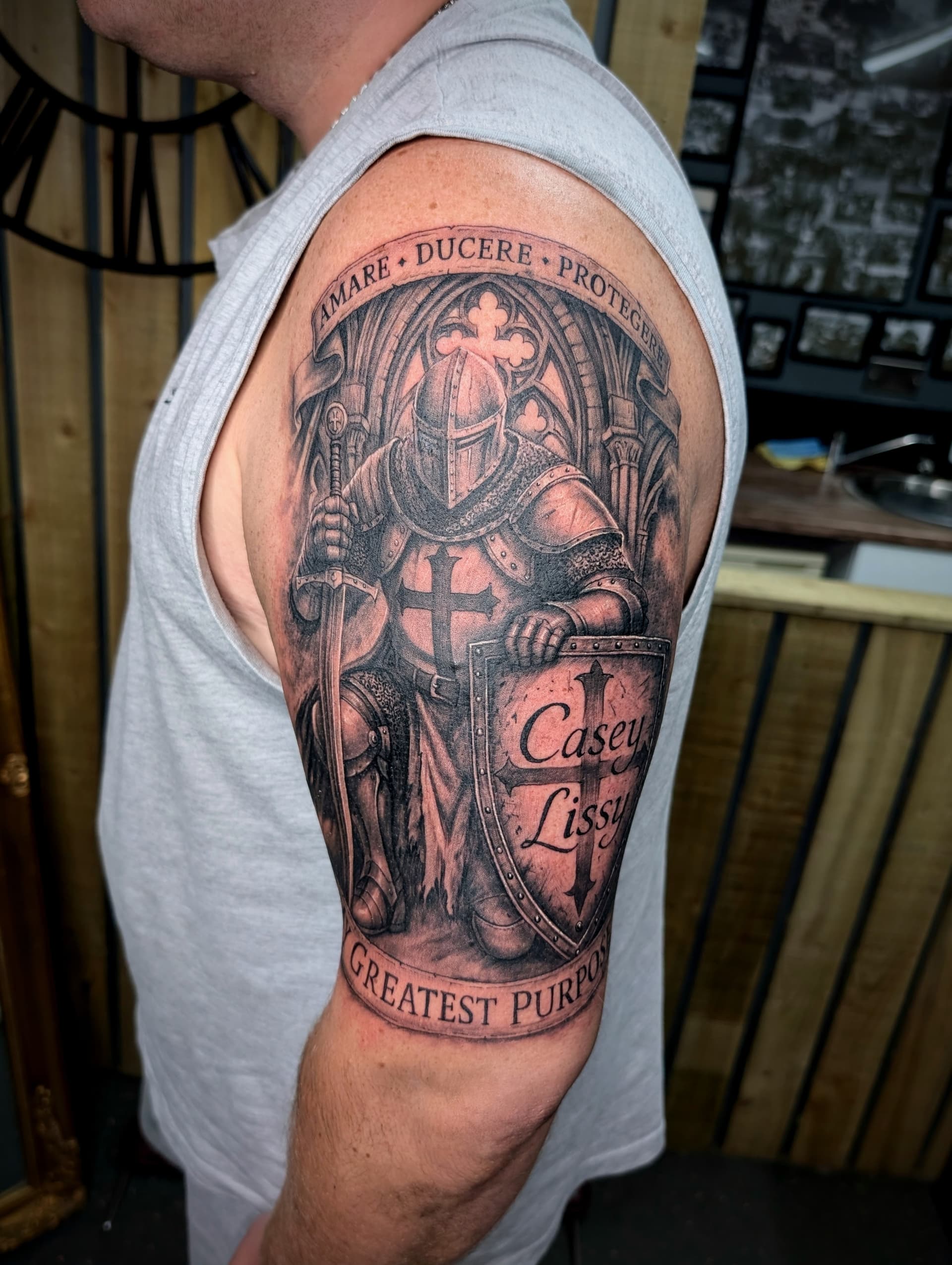 Black and grey realism tattoo of a crusader knight holding a sword and shield with lettering on the upper arm