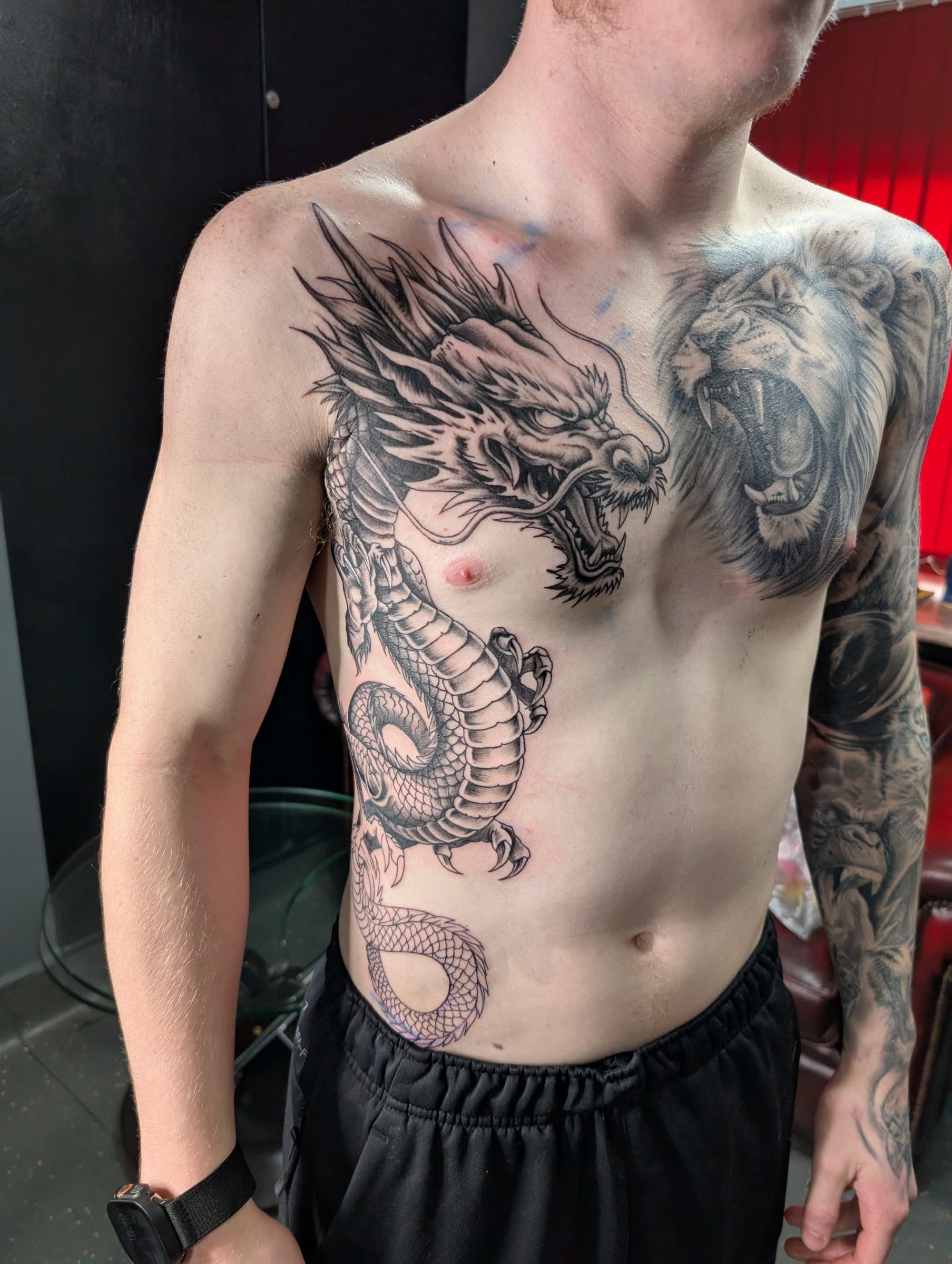 Black and grey realism tattoo of a dragon across the chest and ribs beside a roaring lion on the chest