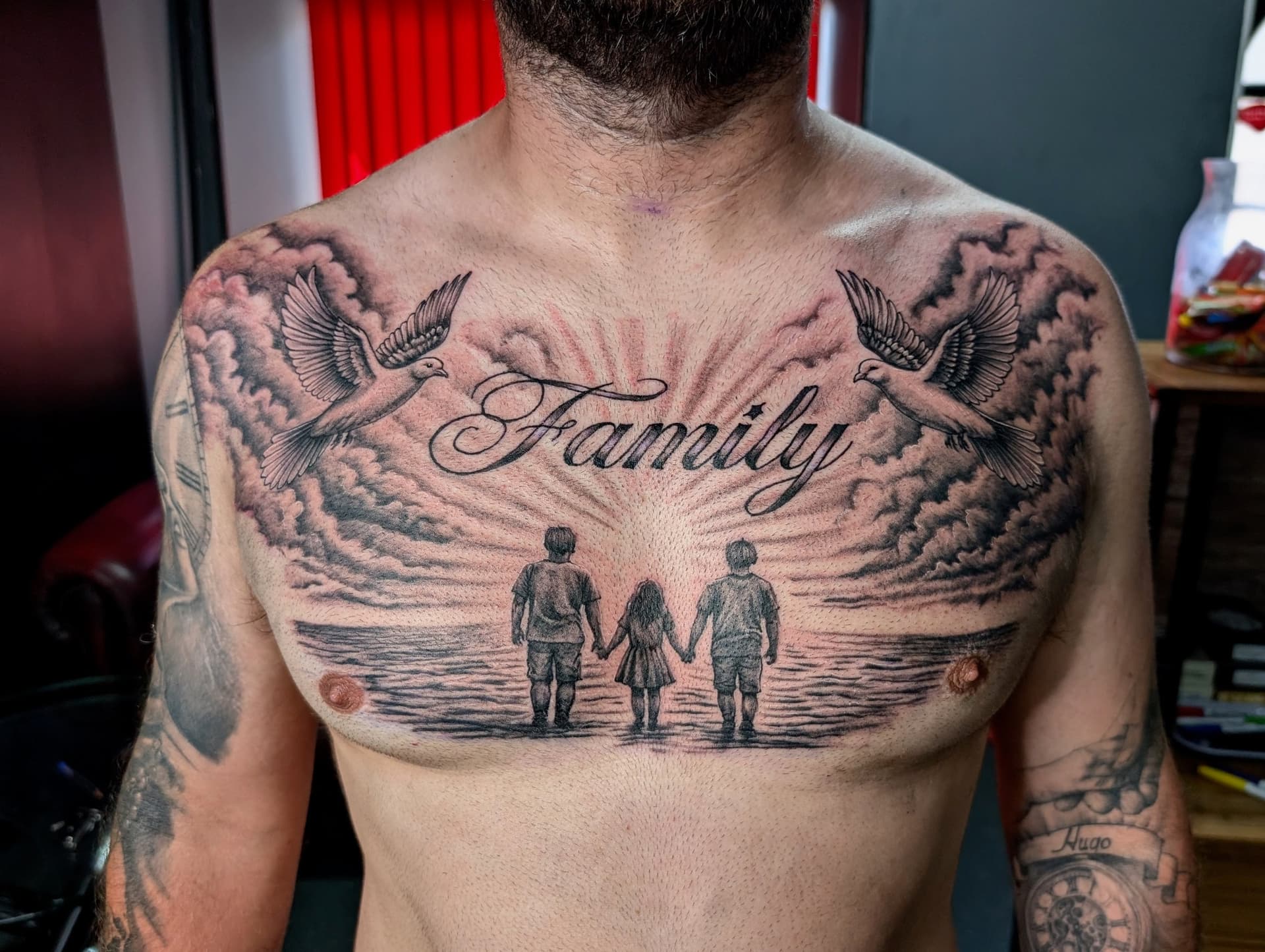 Black and grey realism chest tattoo of three silhouettes walking on a beach with doves and 'Family' script