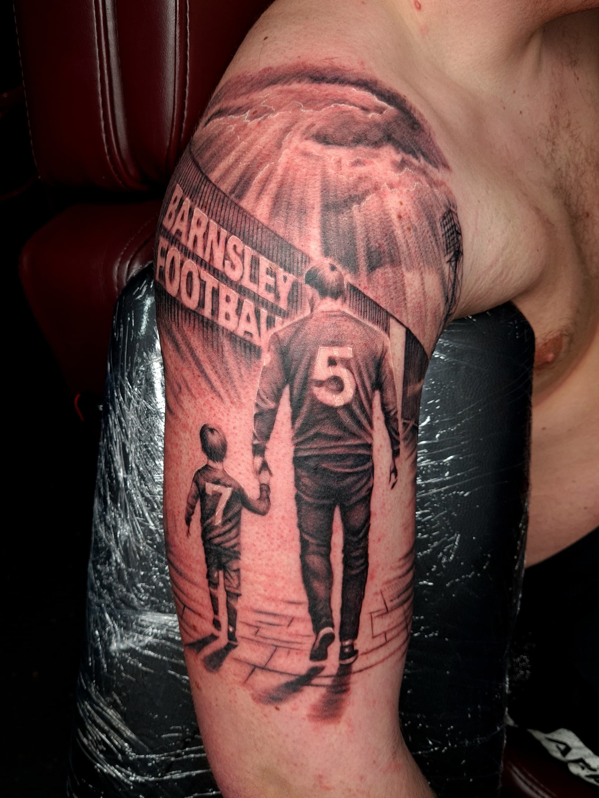 Black and grey realism tattoo of a father and son walking to a football match on the upper arm