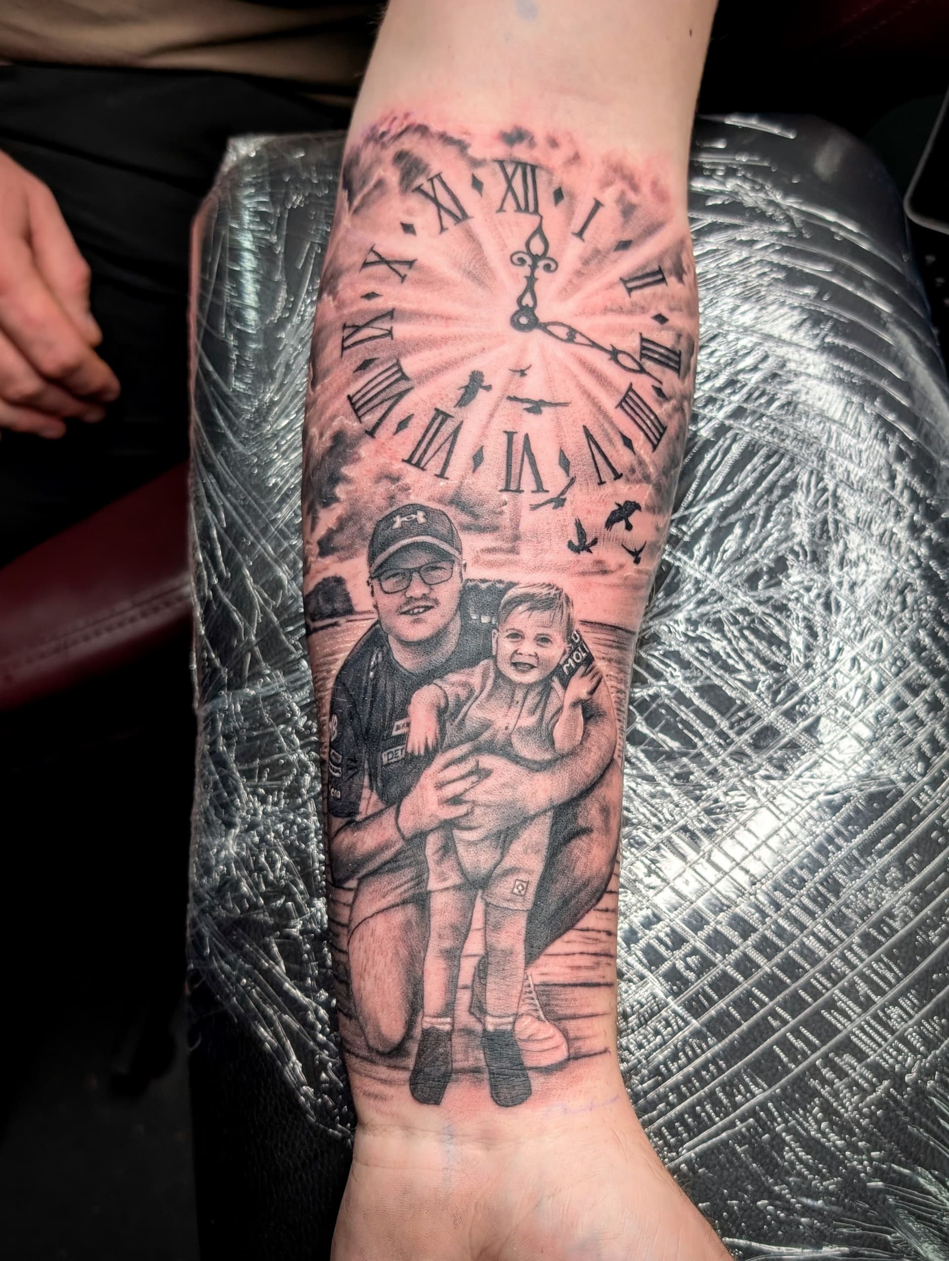 Black and grey realism tattoo of a man holding a child beneath a clock face on the forearm