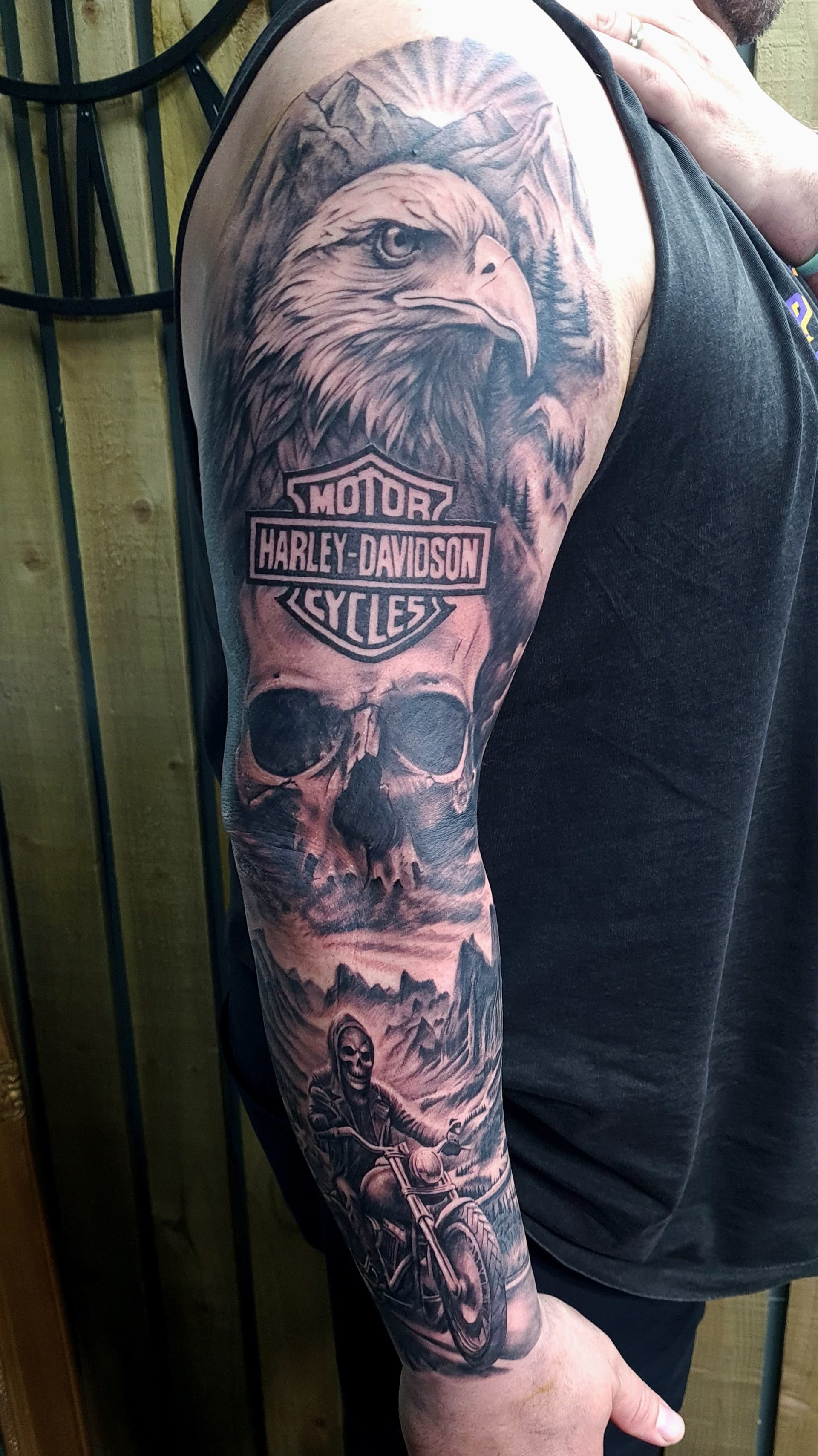 Black and grey realism arm sleeve tattoo of a bald eagle, skull, Harley-Davidson logo and a grim reaper on a motorcycle