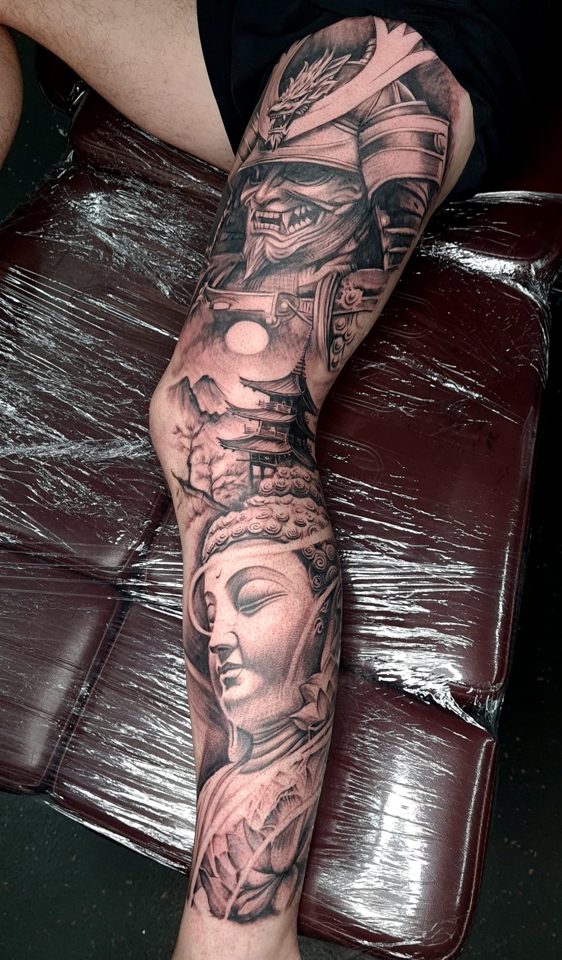 Black and grey realism tattoo of a samurai, Buddha, dragon and pagoda on the leg