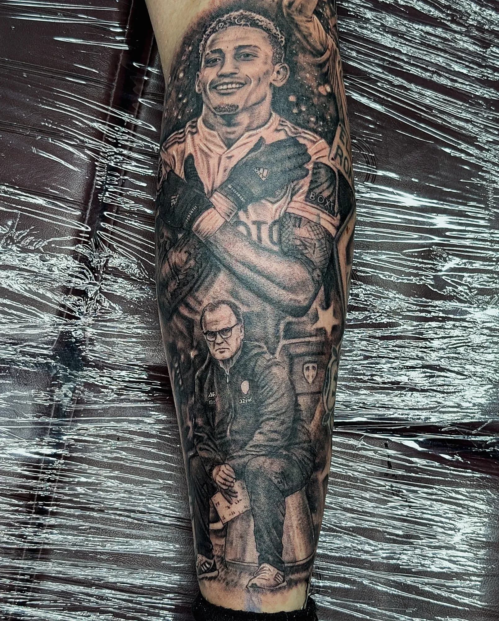 Black and grey realism tattoo of a Leeds United player celebrating above a seated manager, on the back of a lower leg.