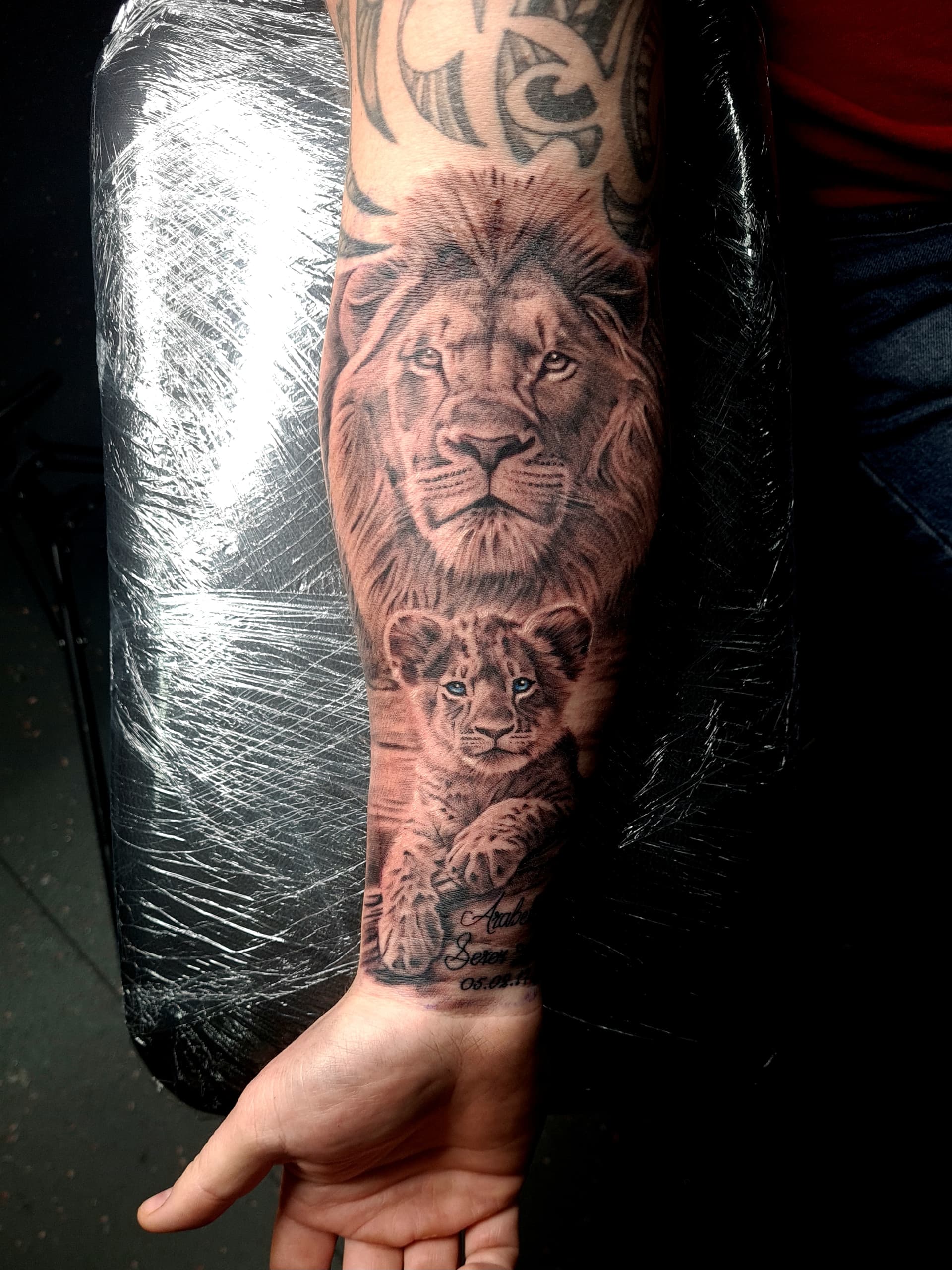 Black and grey realism tattoo of a lion and lion cub with names in script on the forearm