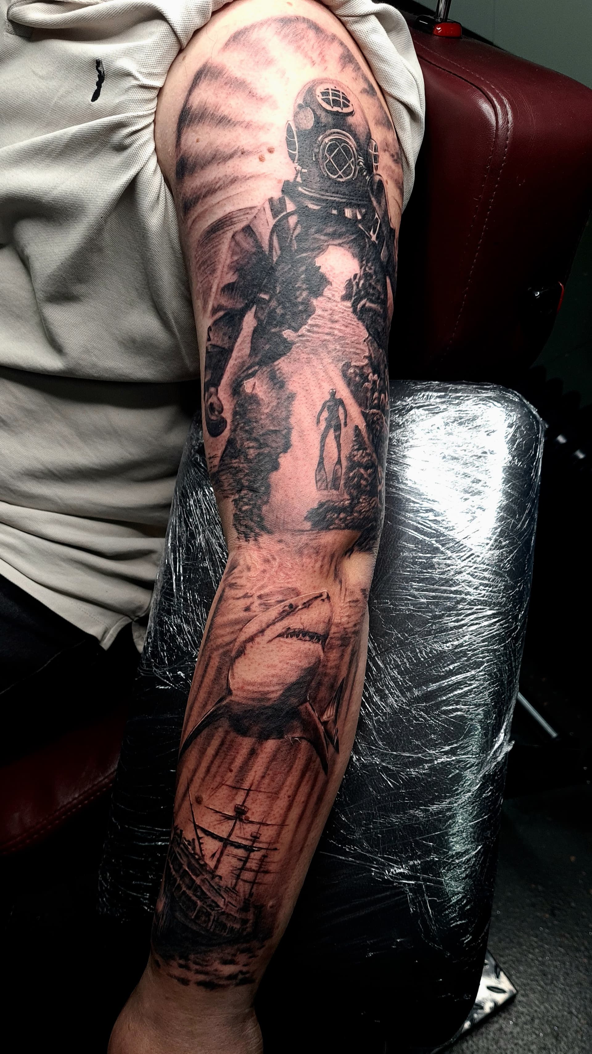 Black and grey realism arm sleeve tattoo featuring a diving helmet, diver, shark and sailing ship