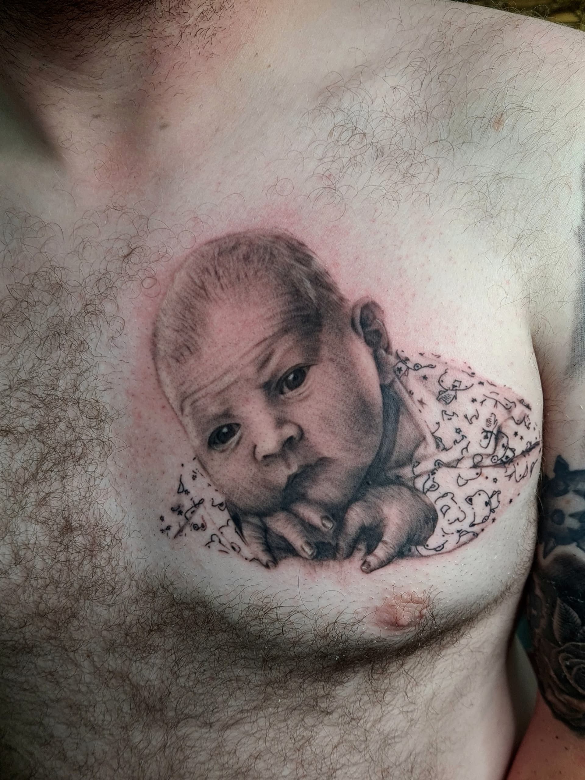 Black and grey realism tattoo of a newborn baby wrapped in a blanket on the chest