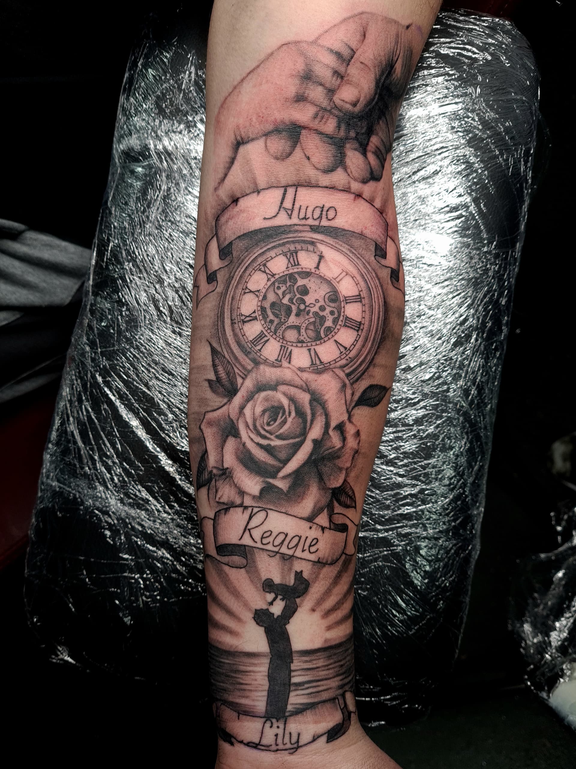 Black and grey realism tattoo of a pocket watch, rose and name banners on the forearm