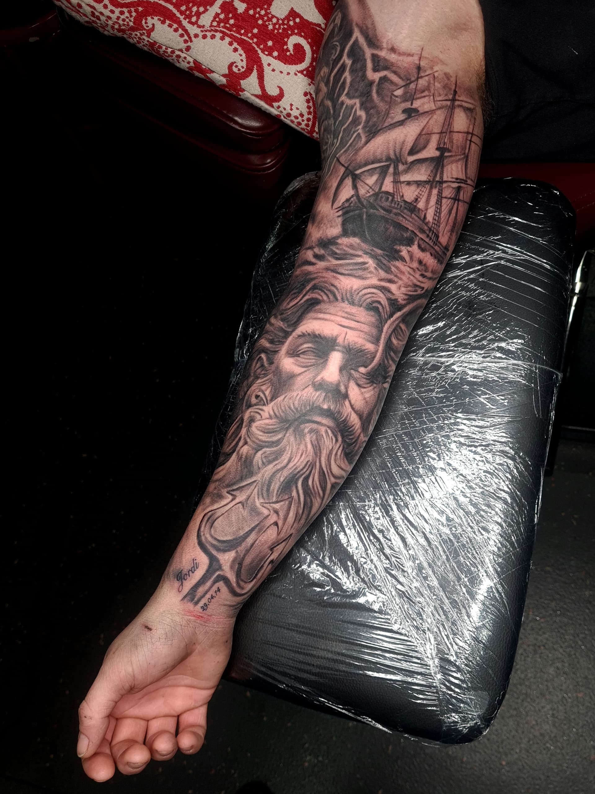 Black and grey realism tattoo of Poseidon, a trident and a sailing ship on the forearm