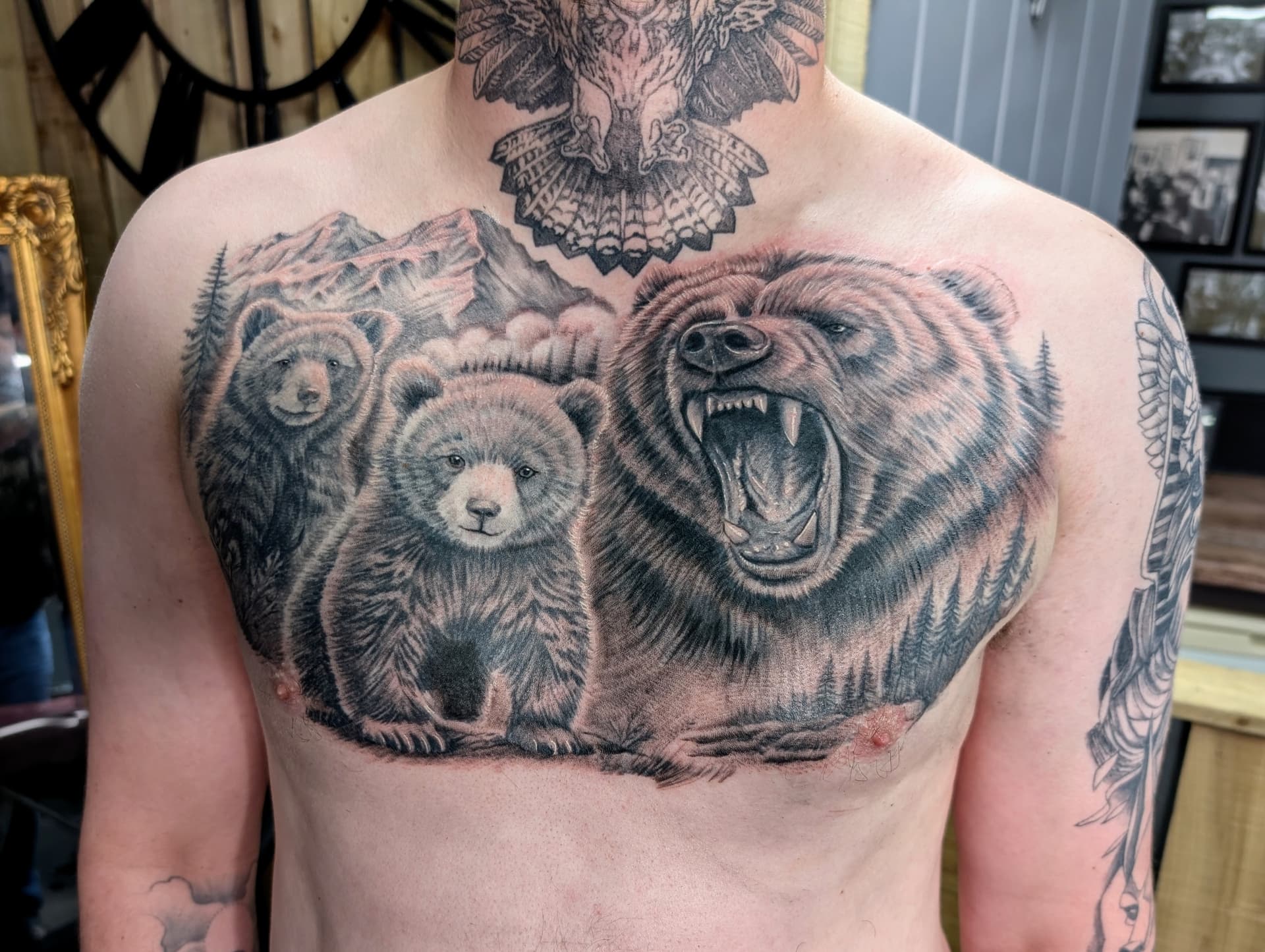 Black and grey realism tattoo of a roaring grizzly bear with two cubs and mountain scenery on the chest