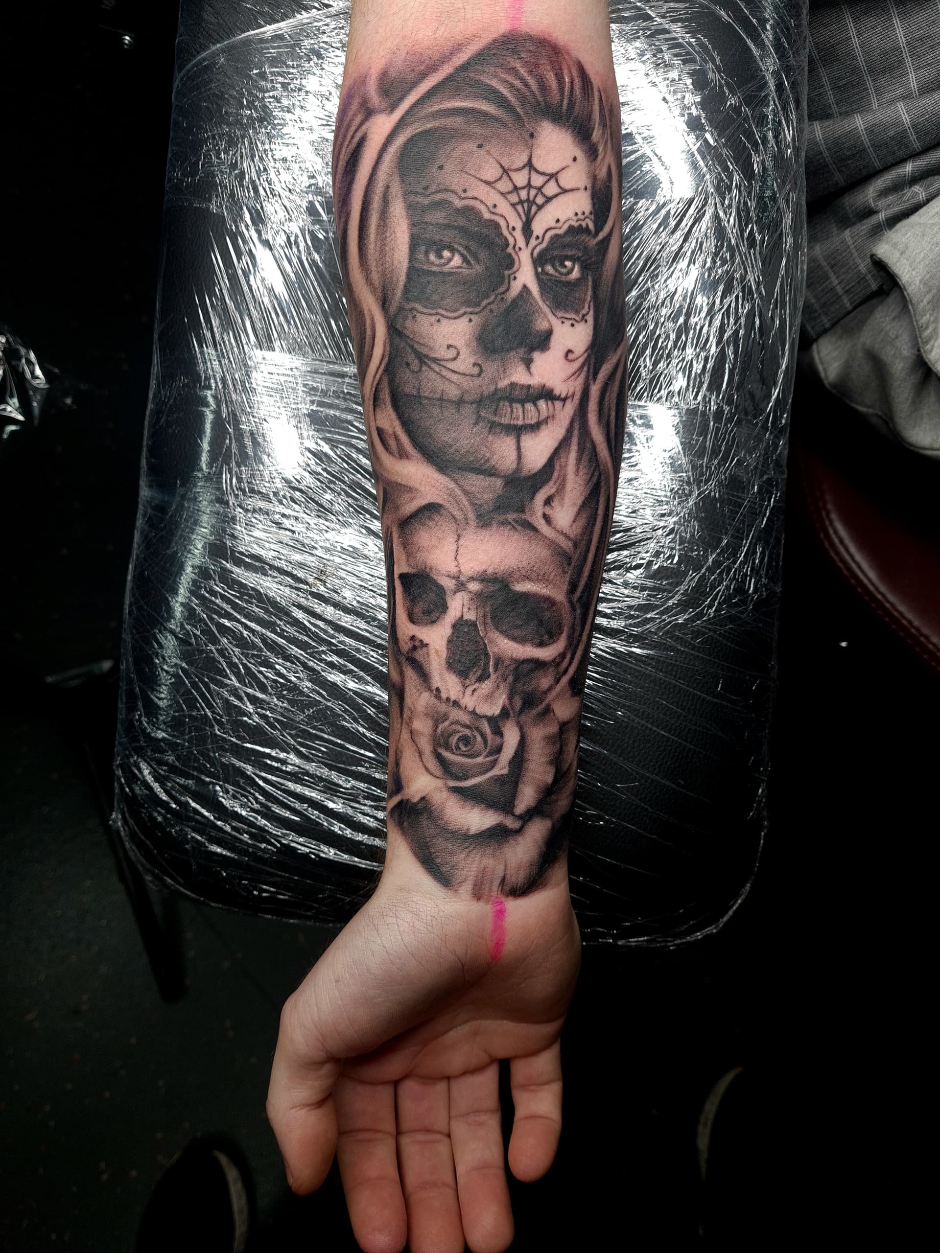 Black and grey realism tattoo of a sugar skull-faced woman, skull and rose on the forearm