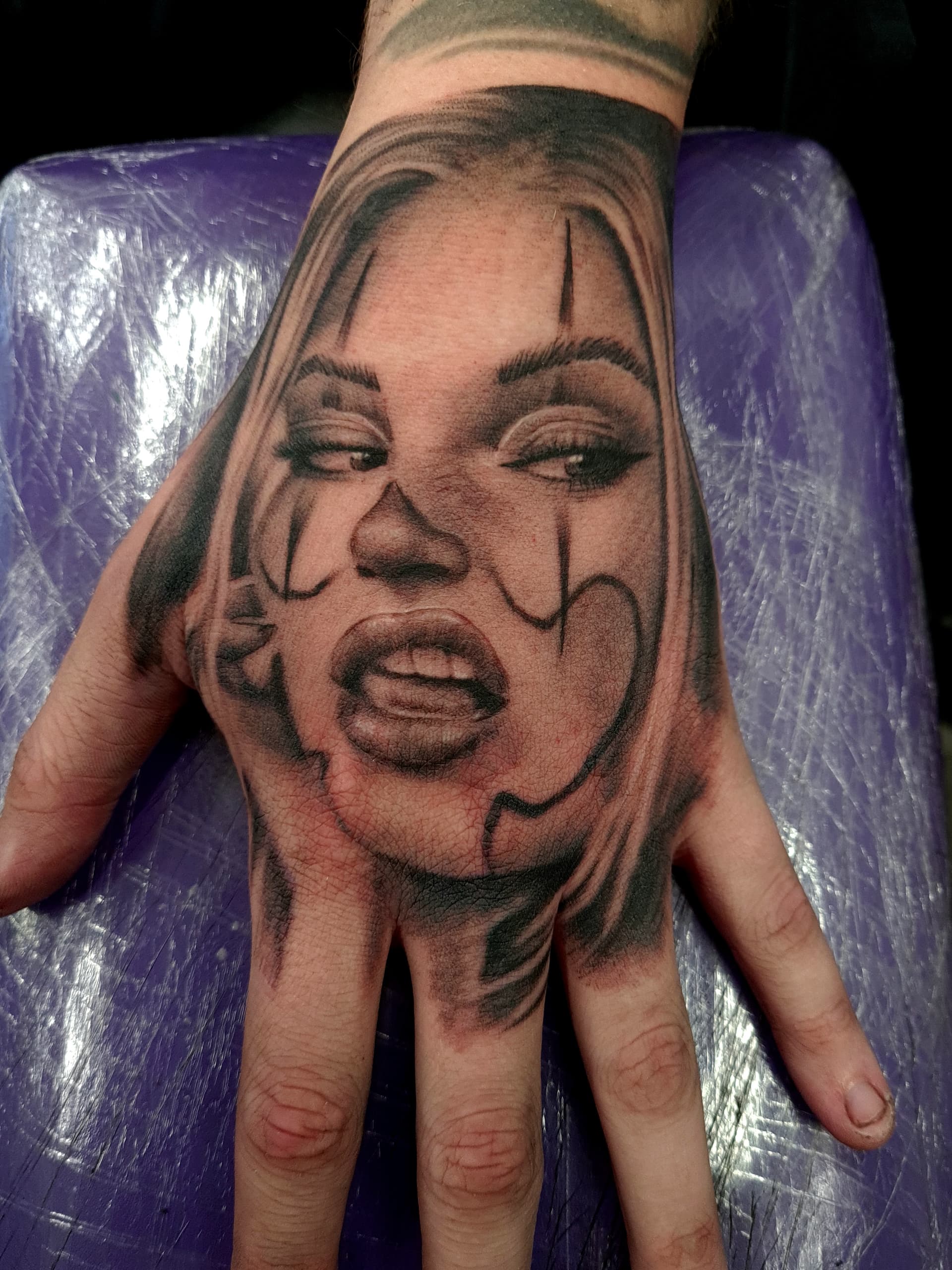 Black and grey realism tattoo of a woman's face with clown makeup on the back of the hand