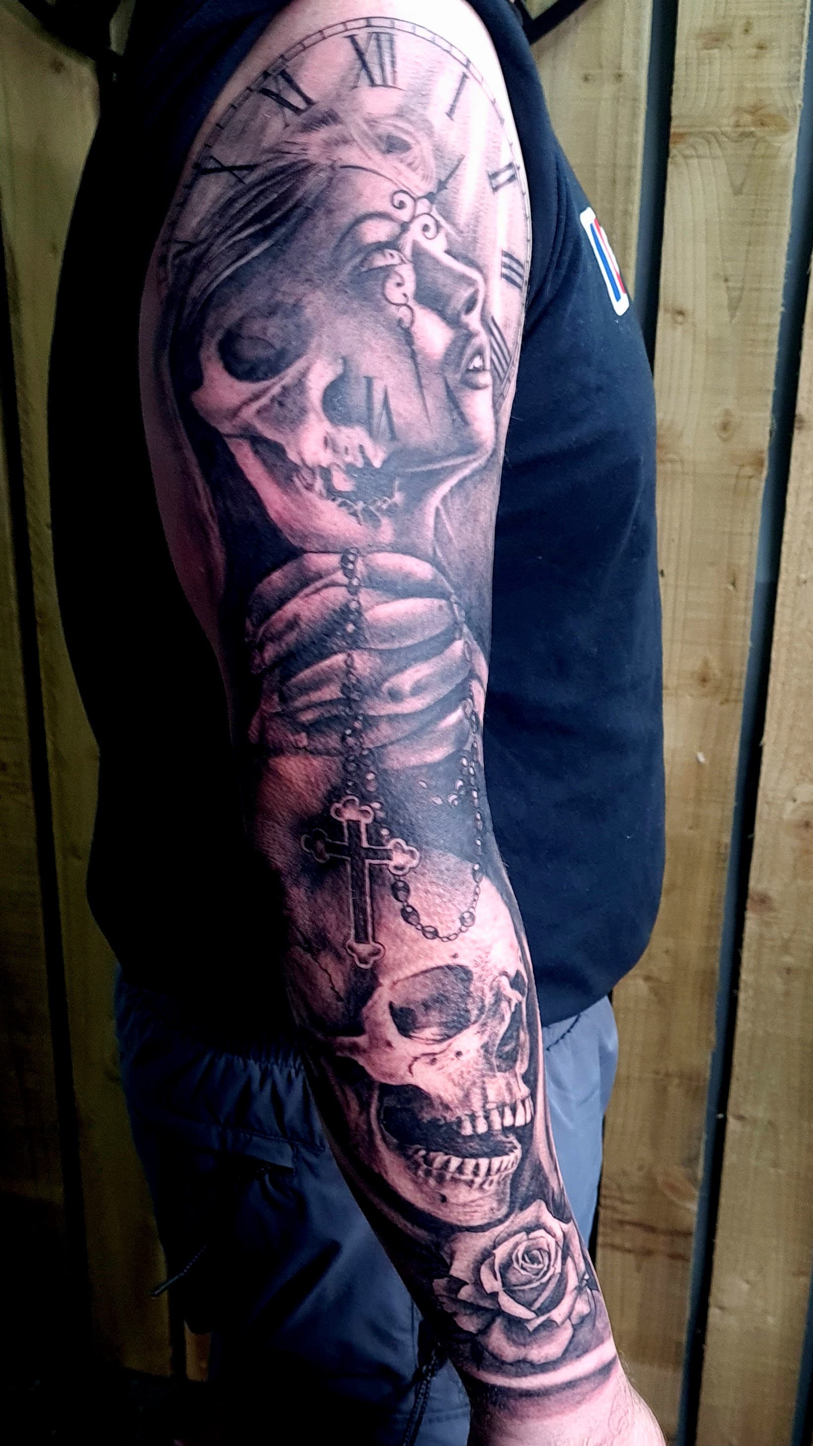Black and grey realism tattoo of a half-skull woman, clock face, rosary and rose on the upper arm