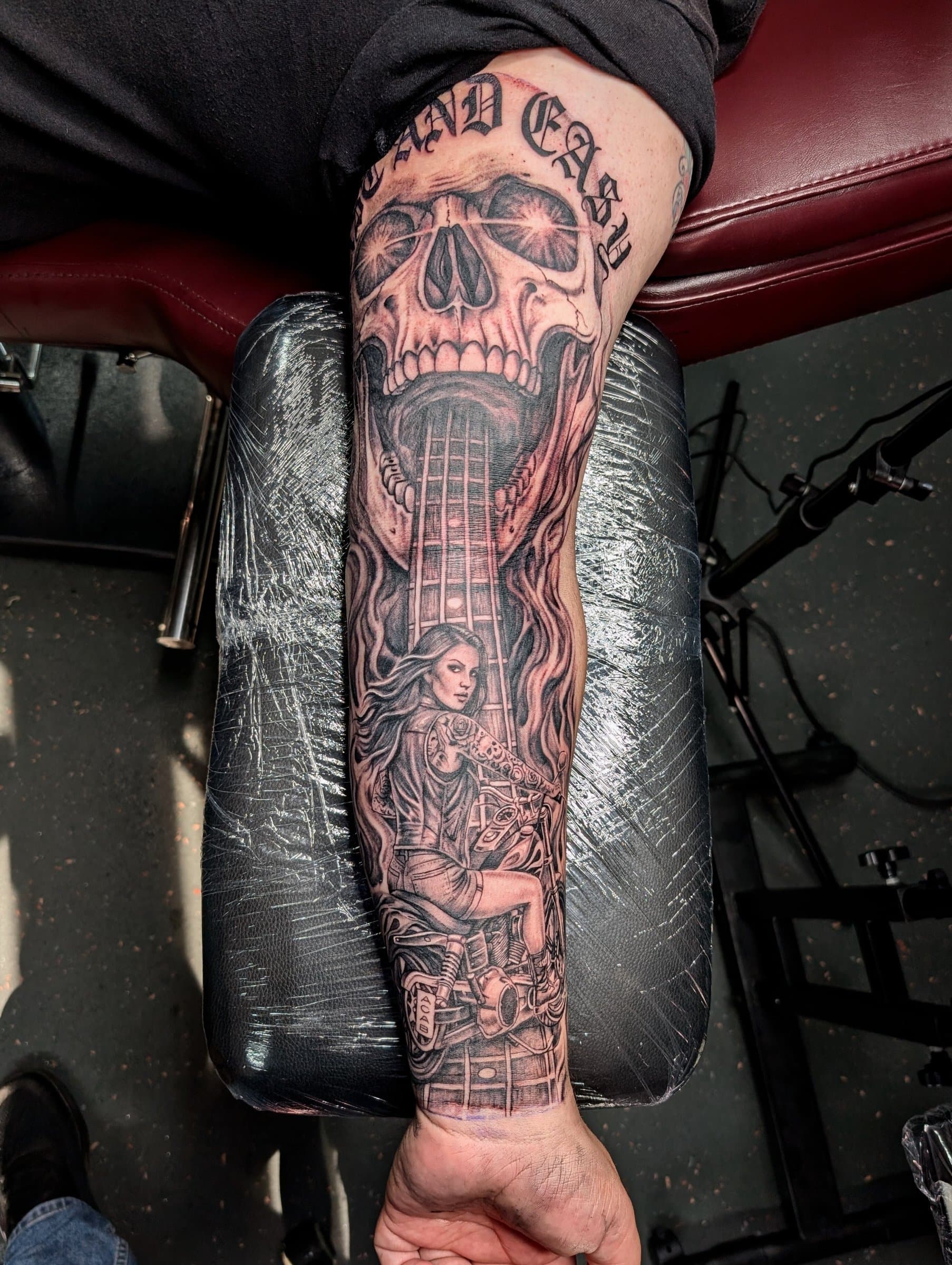 Black and grey realism forearm tattoo of a screaming skull, guitar neck and woman riding a motorcycle