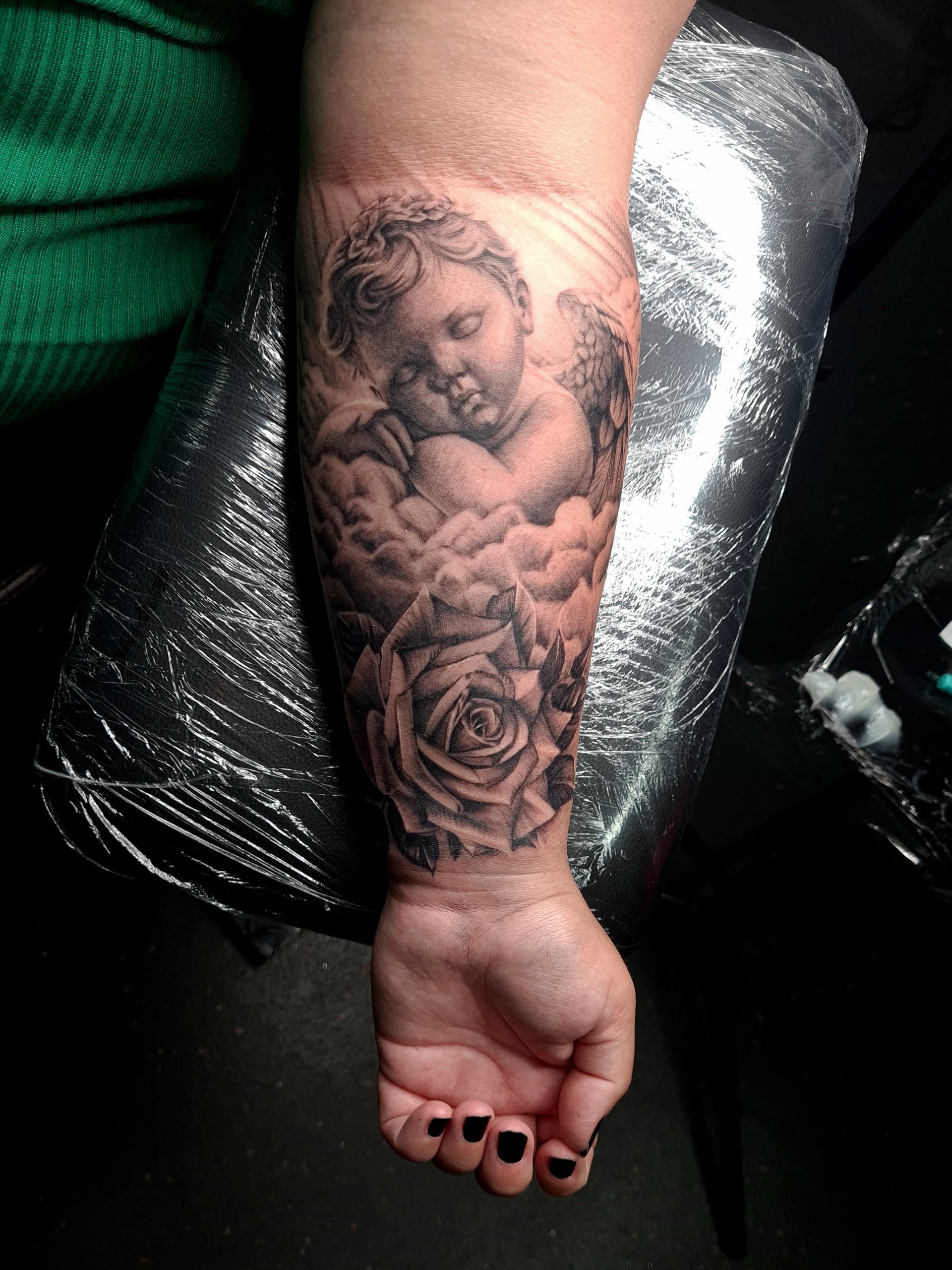 Black and grey realism tattoo of a sleeping cherub angel among clouds and a rose on the forearm