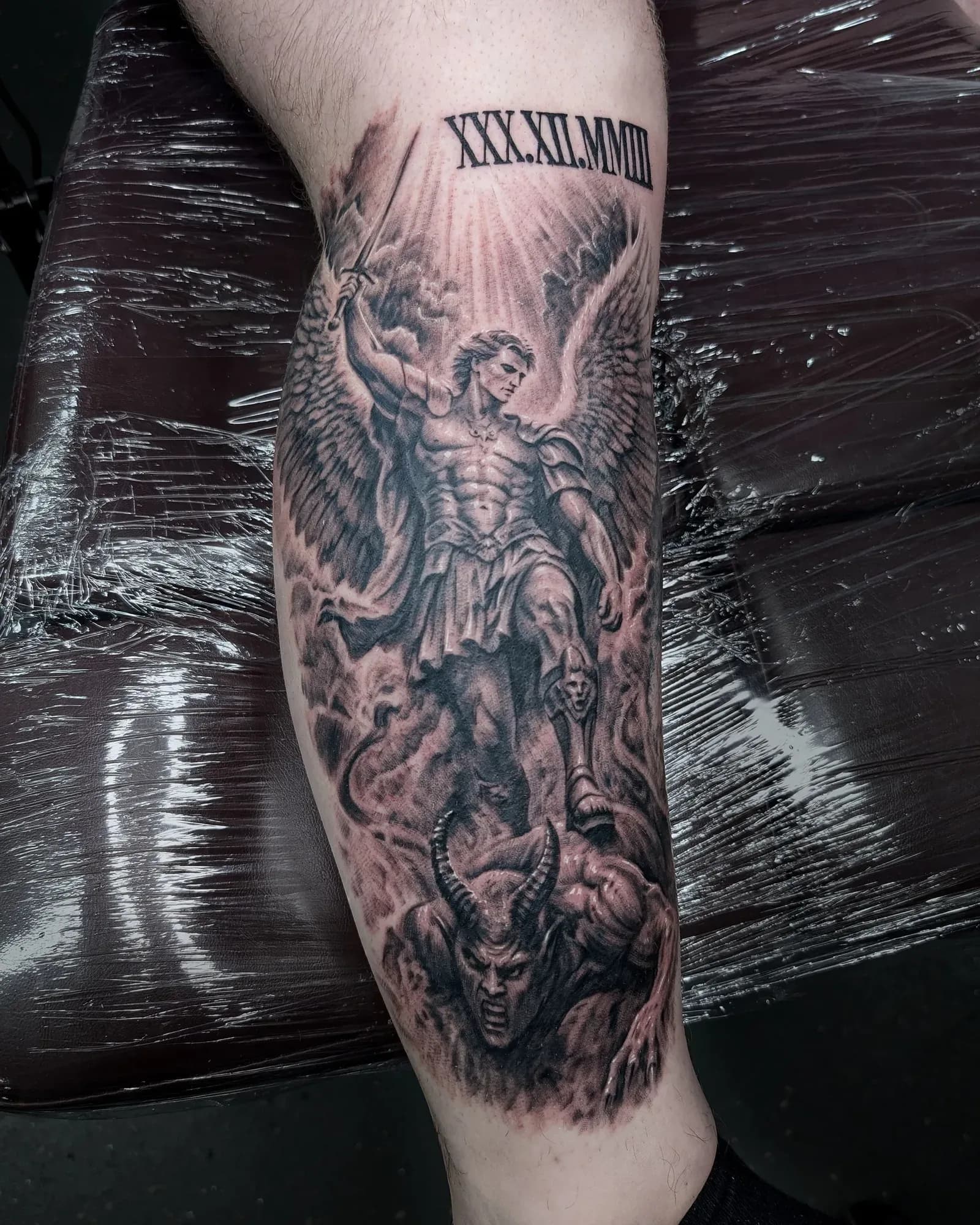 Black and grey realism tattoo of St Michael standing over the devil with his sword, clouds with light breaking through, a