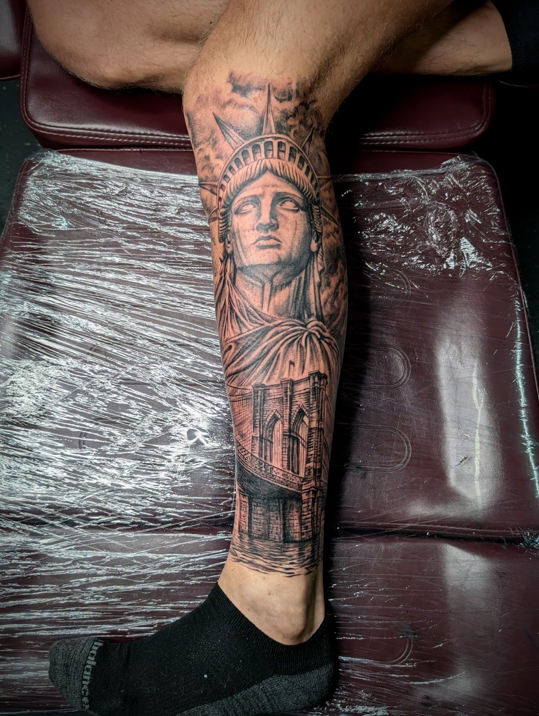 Black and grey realism tattoo of the Statue of Liberty and Brooklyn Bridge on the calf and shin