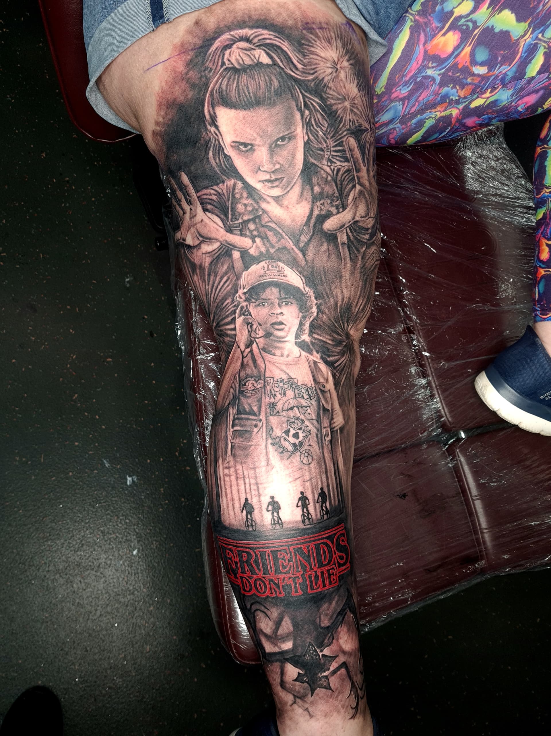 Black and grey realism tattoo of Eleven and Dustin from Stranger Things on the thigh with 'Friends Don't Lie' script