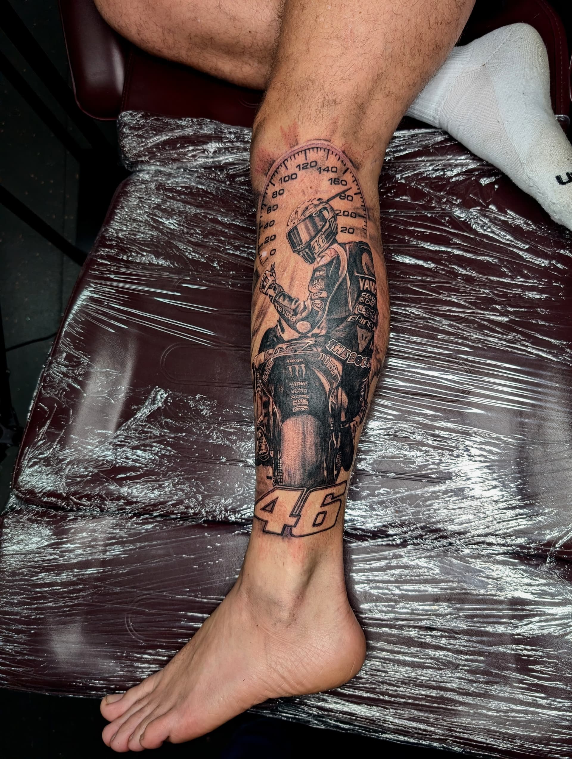 Black and grey realism tattoo of motorbike racer Valentino Rossi with speedometer dial and number 46 on the calf