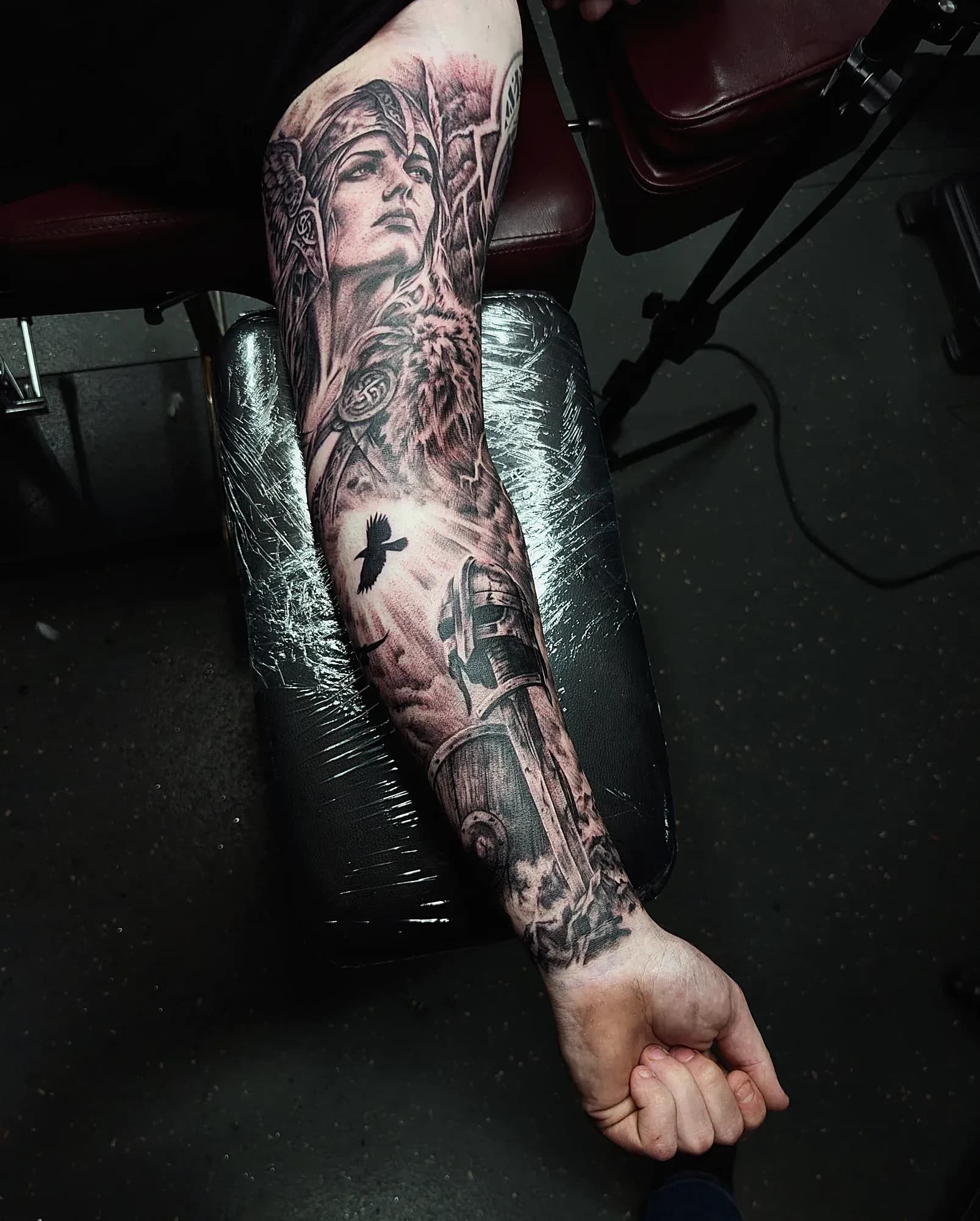 Black and grey realism tattoo of a Norse valkyrie above a fallen Viking warrior and raven, covering the inner arm.