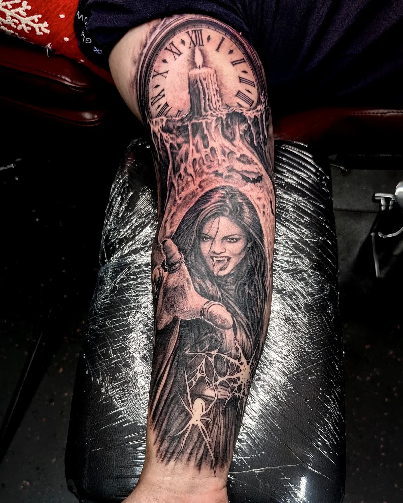 Black and grey realism tattoo of a fanged woman, candle, clock face and spider web running from upper arm to wrist.