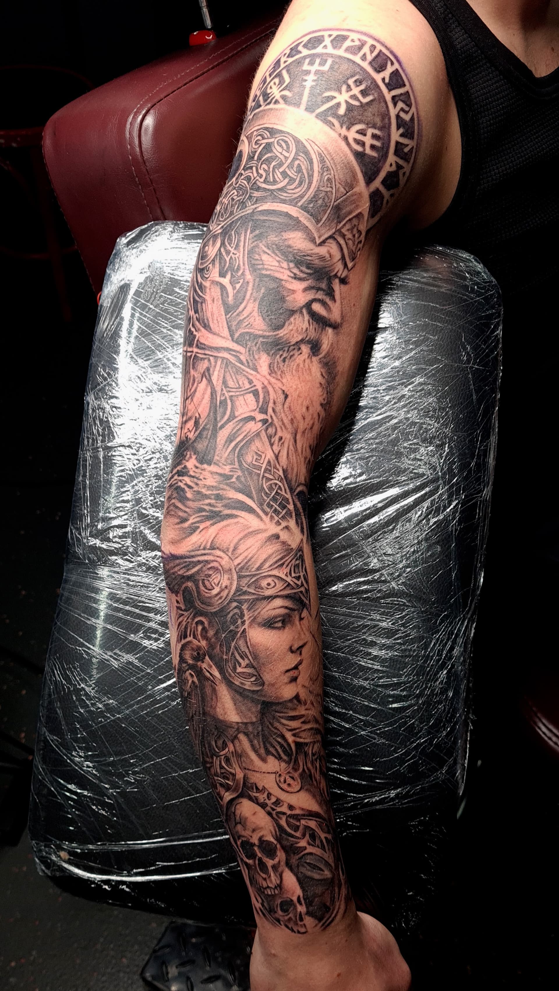 Black and grey realism arm sleeve tattoo featuring Odin, a Viking warrior woman, runes and a skull