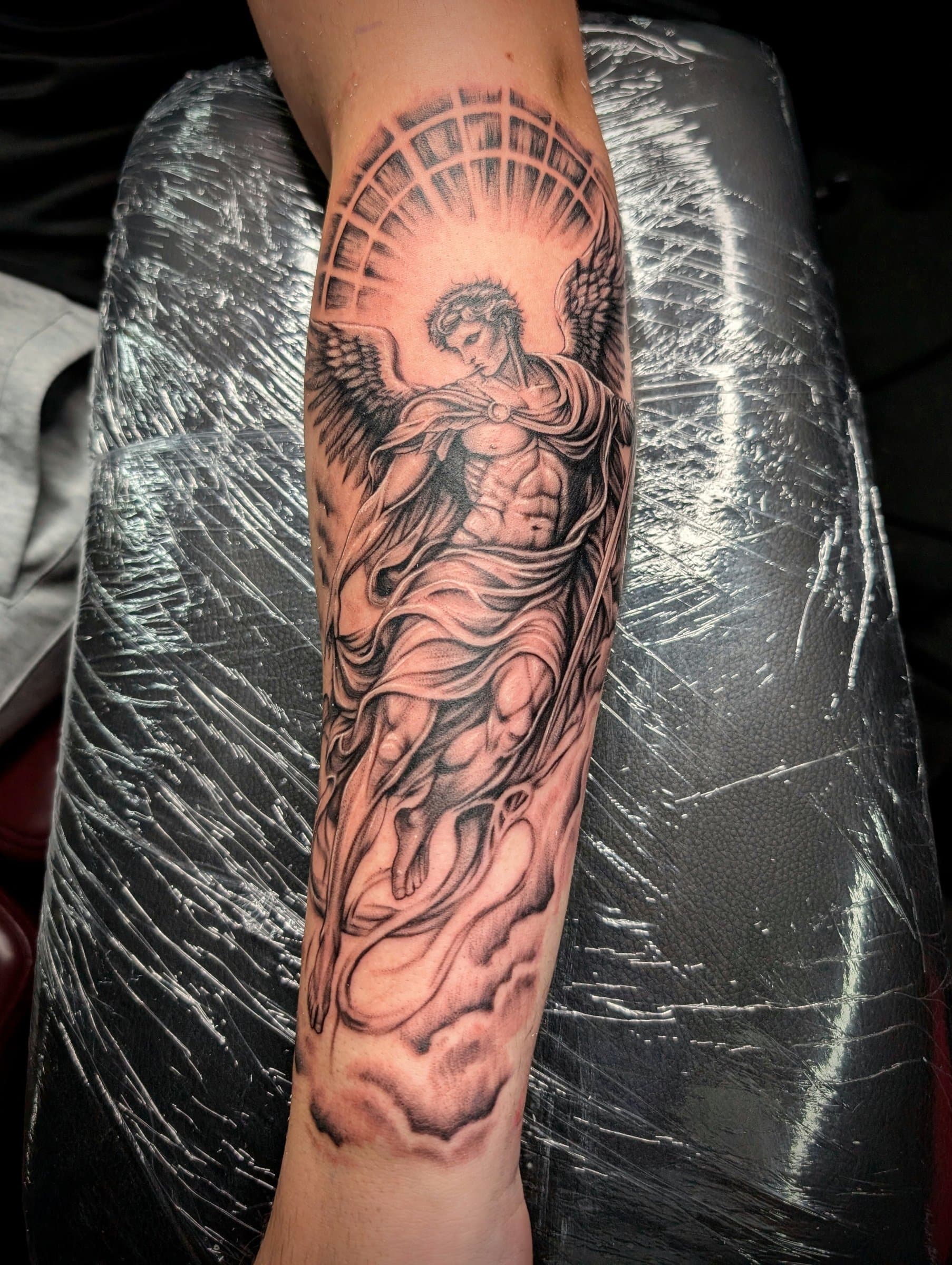 Black and grey realism tattoo of a winged angel with a halo on the forearm