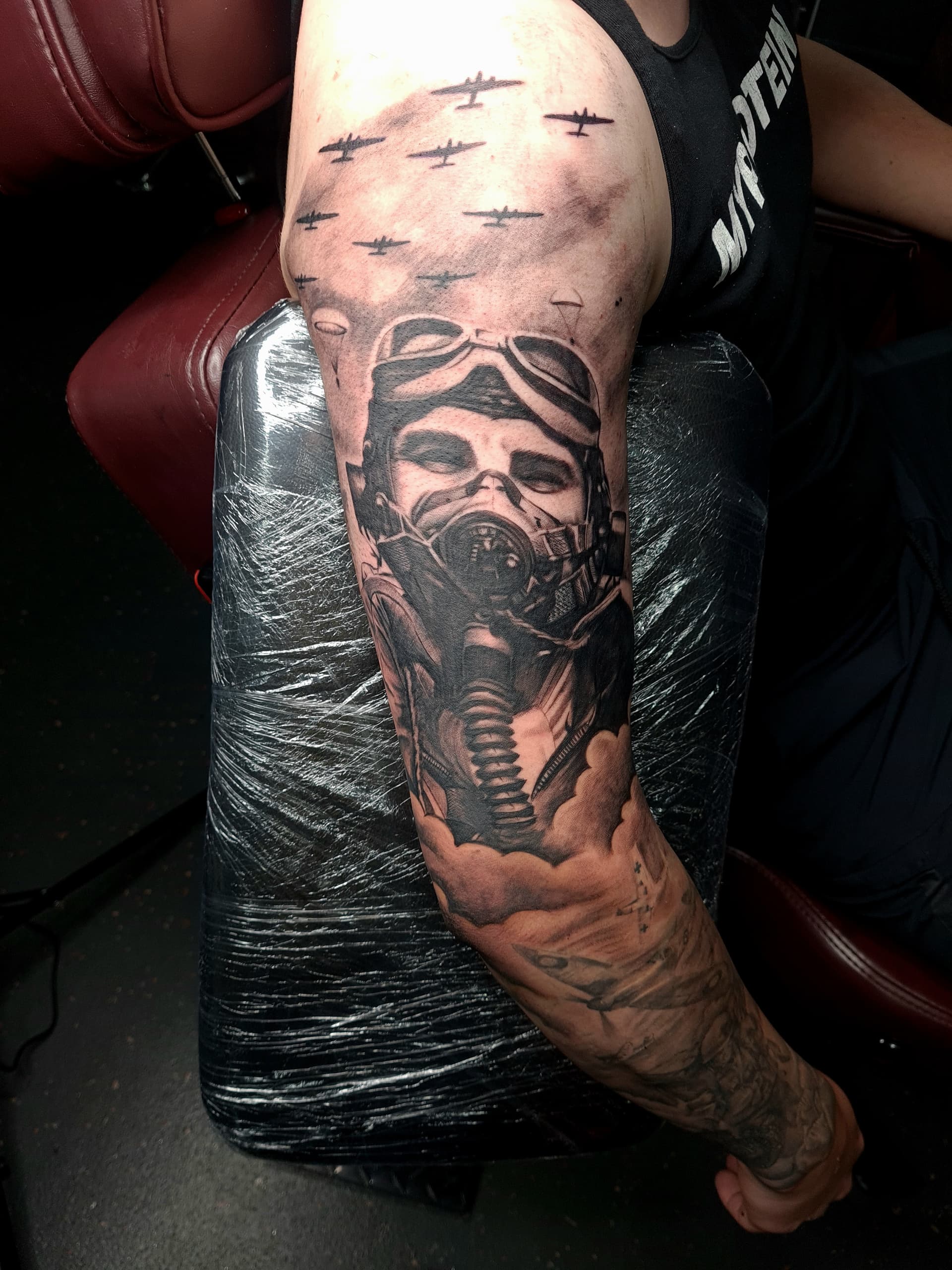 Black and grey realism tattoo of a WWII pilot wearing an oxygen mask beneath a squadron of bomber planes on the inner arm