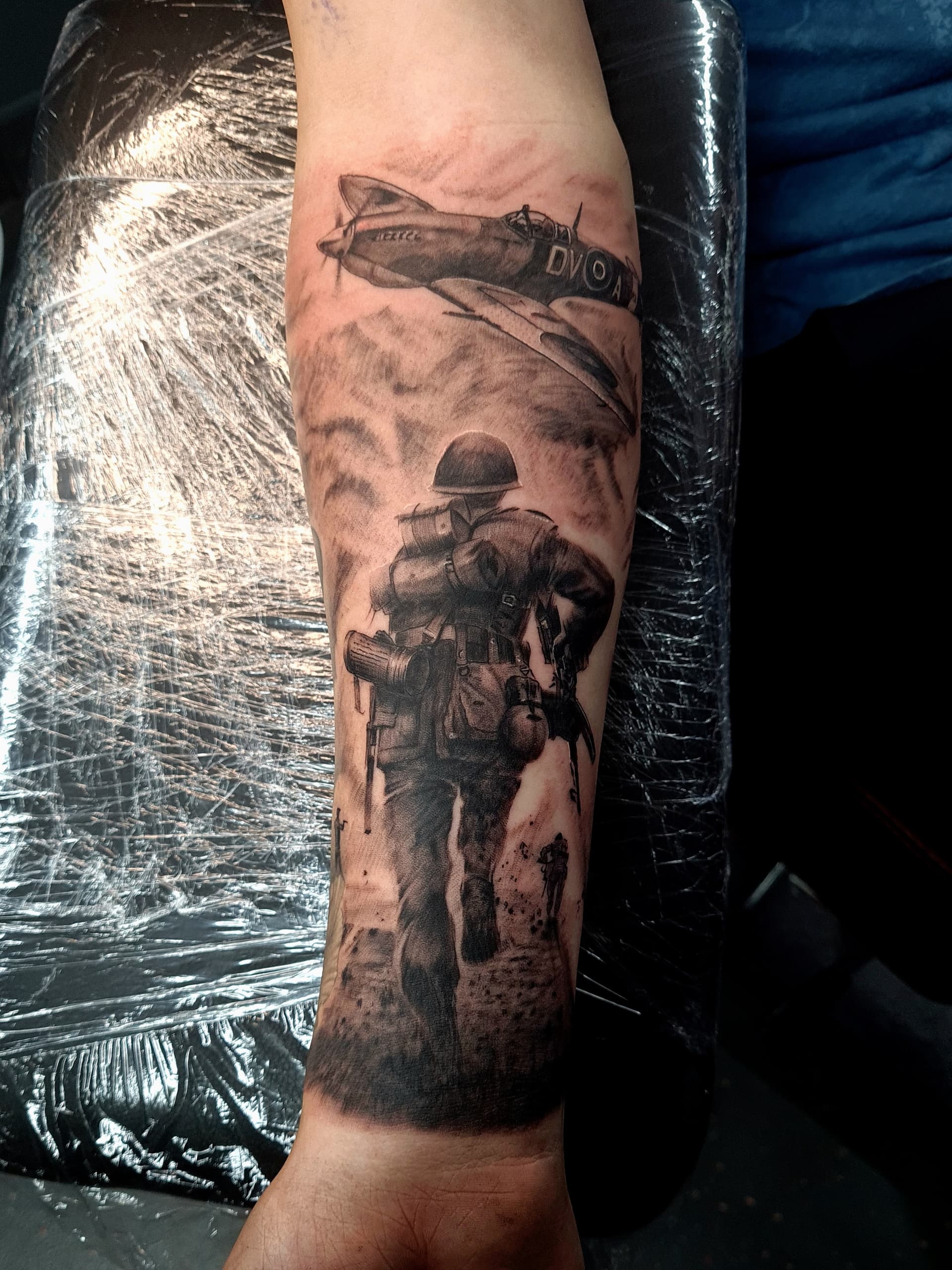 Black and grey realism tattoo of a WWII soldier marching beneath a Spitfire fighter plane on the forearm