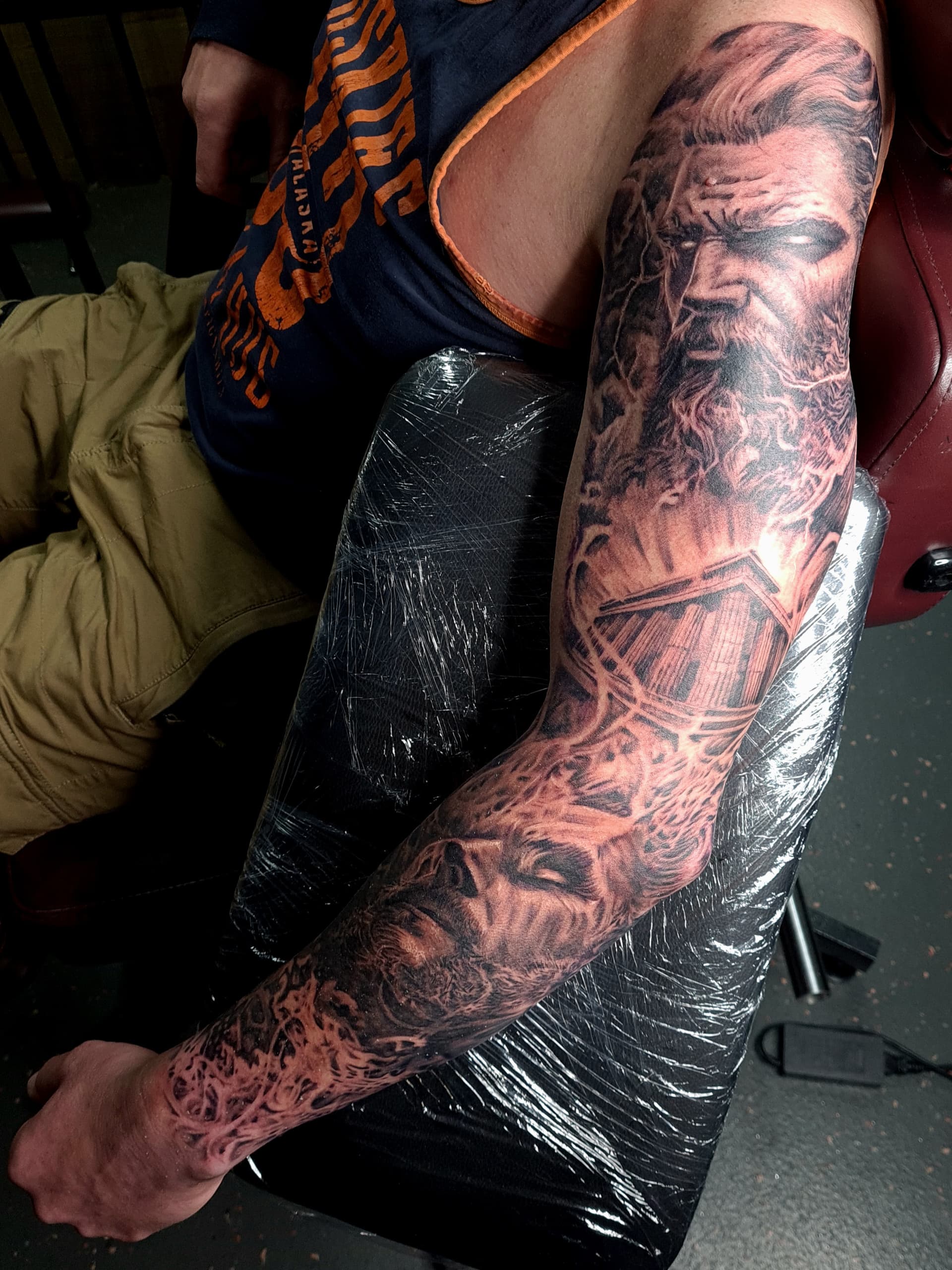 Black and grey realism tattoo of a mythological god Zeus, a cabin and a wolf covering the full arm